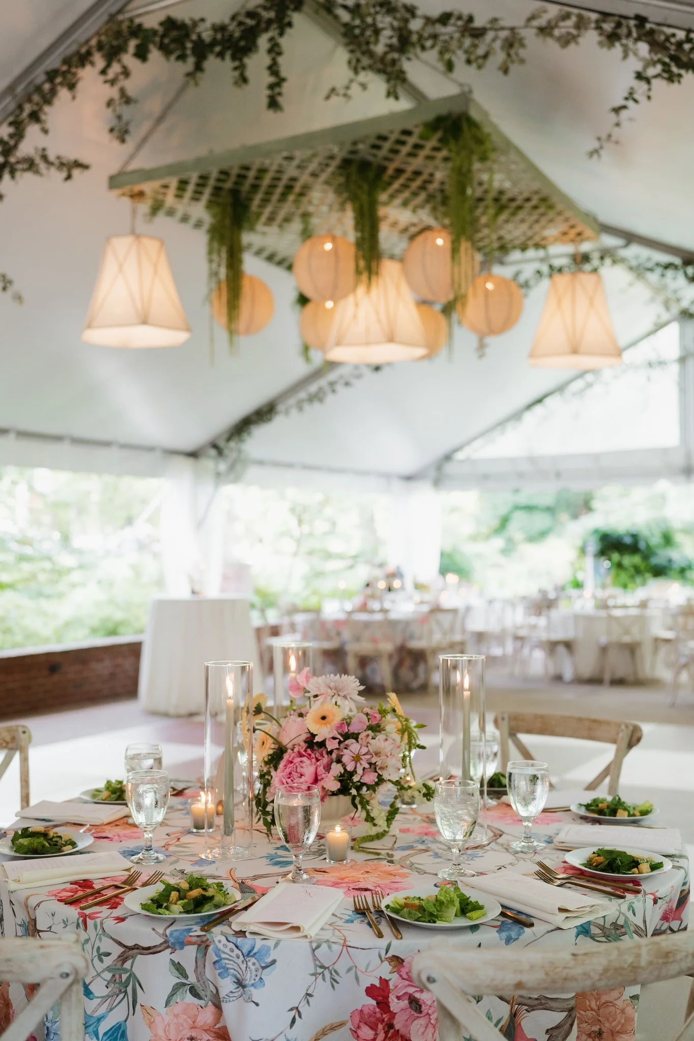 A decorated indoor event space with a table set for a formal meal, featuring a floral centerpiece, candles, and elegant tableware, under a tent covering with hanging lanterns and greenery in Memphis, TN. 