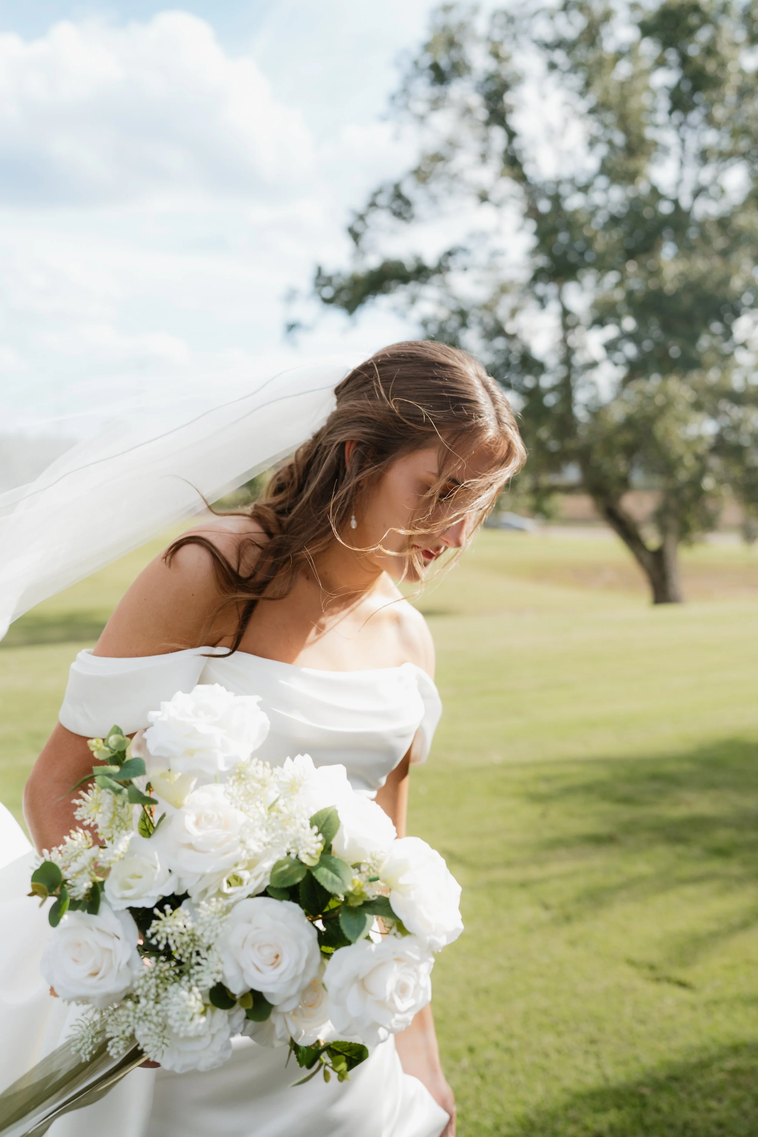 A bride in a white off-shoulder wedding dress holding a bouquet of white roses and greenery, outdoors with green grass and trees, and a blue sky with clouds in the background in Memphis, TN. 