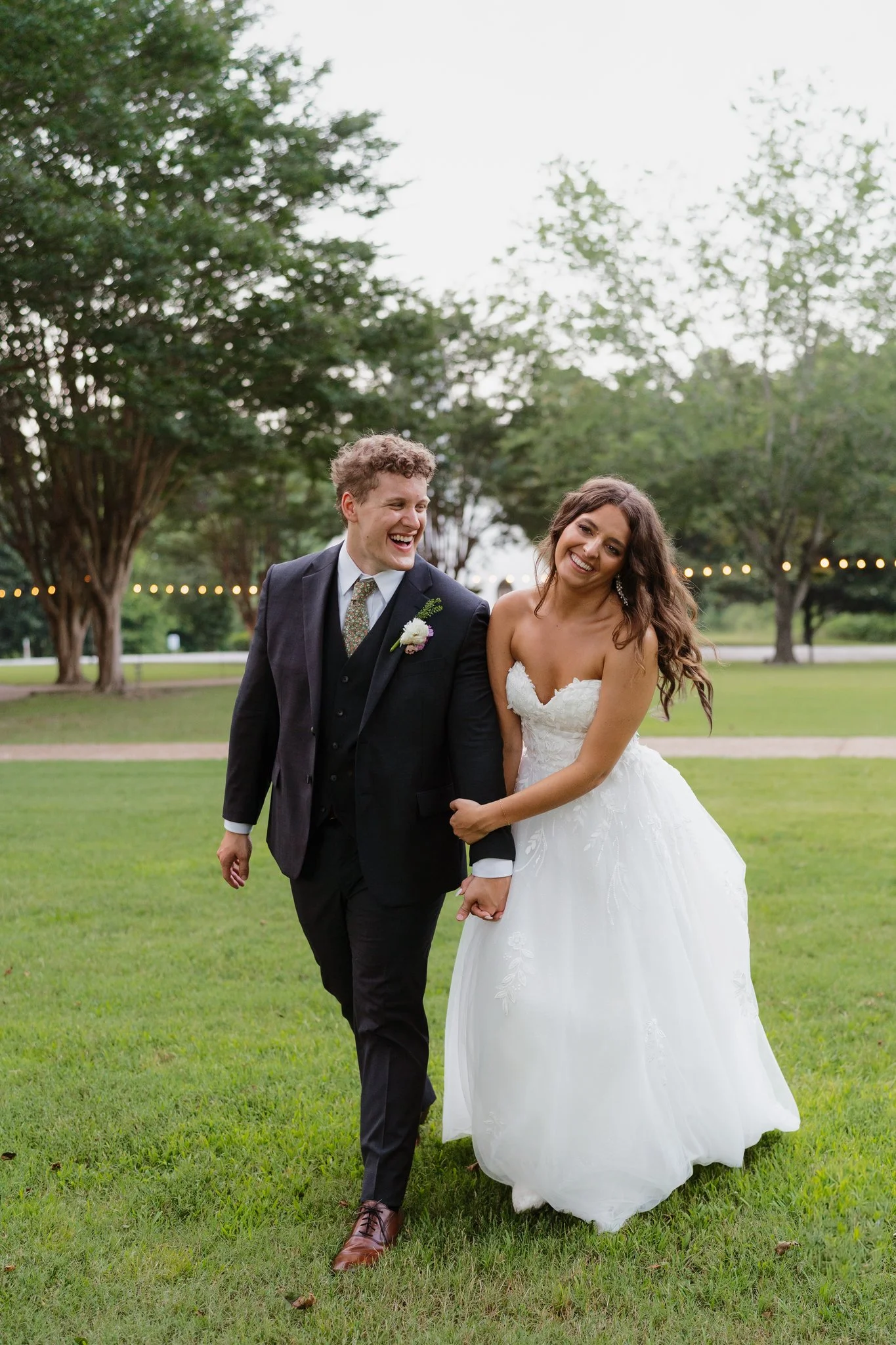 A happy couple, a bride in a white lace wedding dress and a groom in a black suit, holding hands and walking outdoors on a green lawn during a wedding celebration, with trees and string lights in the background in Memphis, TN. 