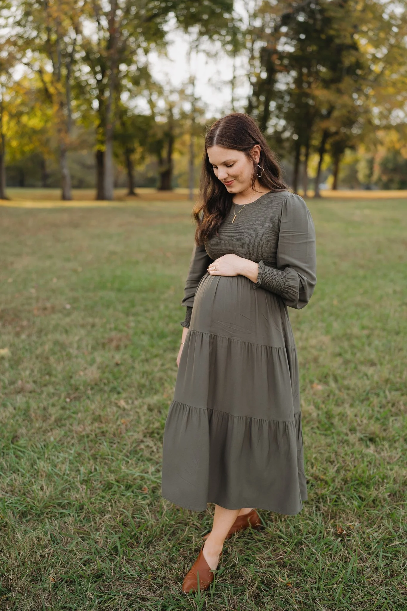 A pregnant woman with long dark hair in a brown dress standing outdoors on grass with trees in the background, gently resting her hand on her belly and smiling down in Memphis, TN. 
