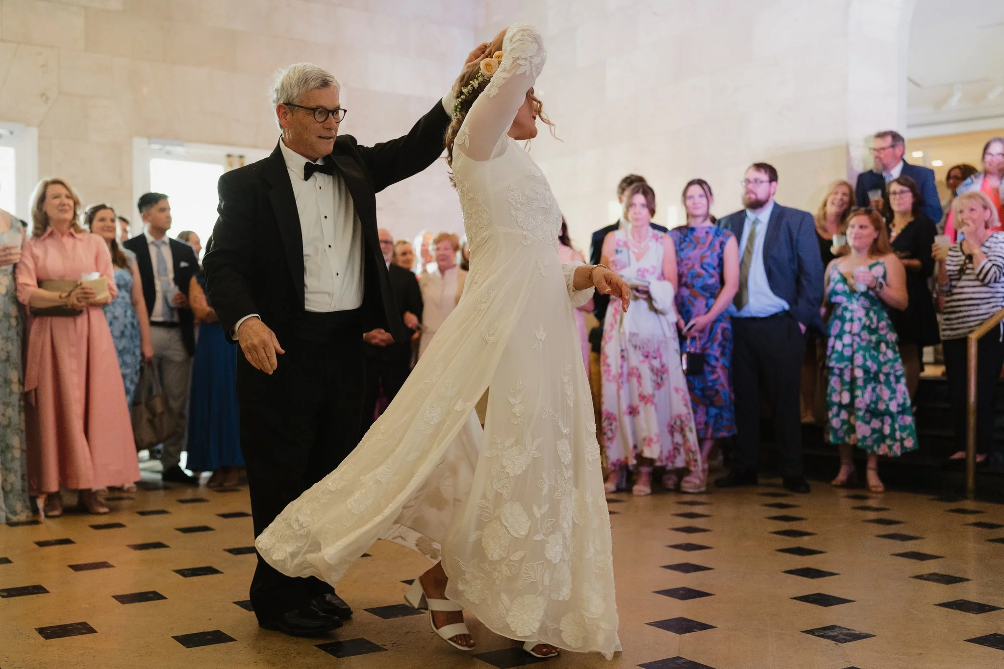 A bride in a white lace wedding dress with floral embroidery lifts her arm as she and her father dance at a wedding reception, surrounded by guests in colorful dresses and suits in Memphis, TN. 