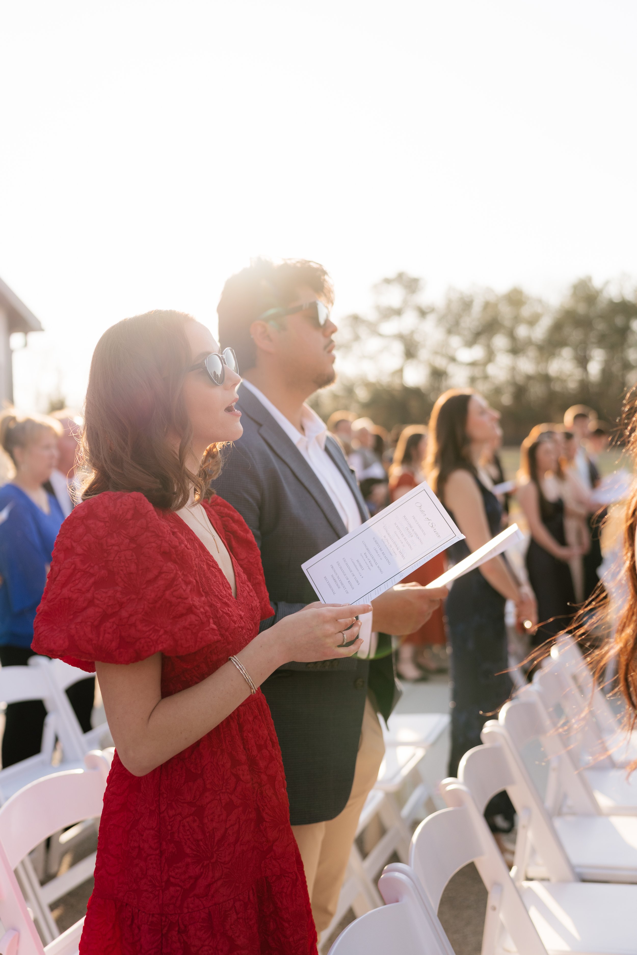A woman in a red dress and a man in a suit with sunglasses are standing outdoors on a sunny day, holding programs, among rows of white chairs, with other people in the background participating in a ceremony in Memphis, TN. 