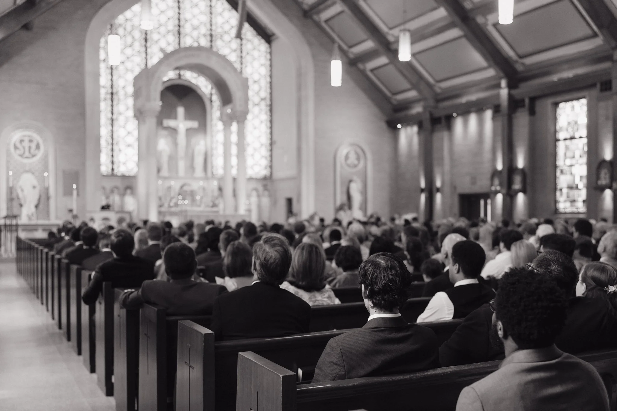 A large congregation of people seated in wooden pews inside a church during a wedding service. The church has high ceilings with hanging lights, stained glass windows, and religious symbols, including a cross at the altar in Memphis, TN. 