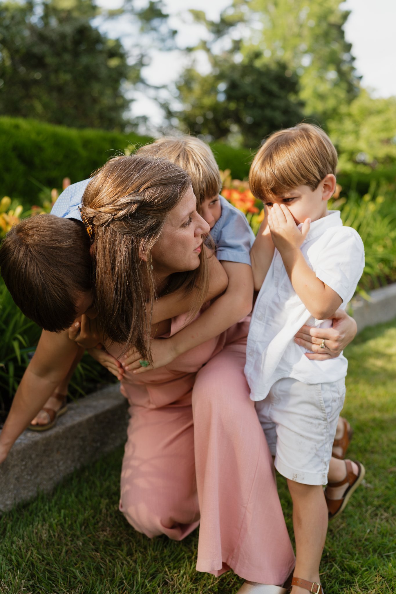 A group of children and a woman are outdoors on a lawn with greenery and flowers in the background. The woman is crouched down with three children around her. One child is in front, covering his nose, and a girl is being held by the woman. The scene 