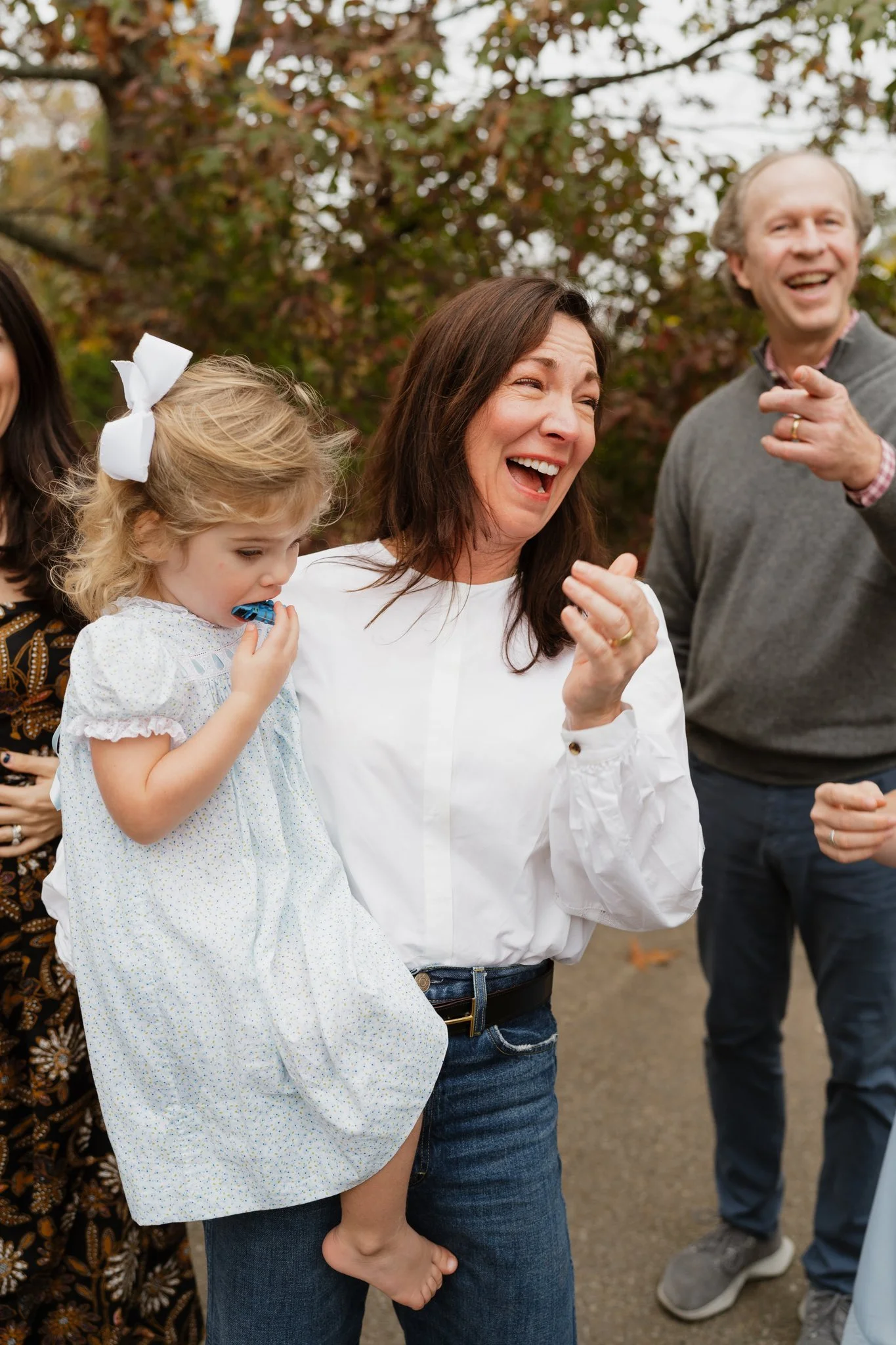 A group of people outdoors, smiling and laughing. A woman with dark hair and a white blouse is in the center, holding a young girl with blonde hair in a white dress with a bow. The girl is eating a blue treat. Other individuals are partially visible 
