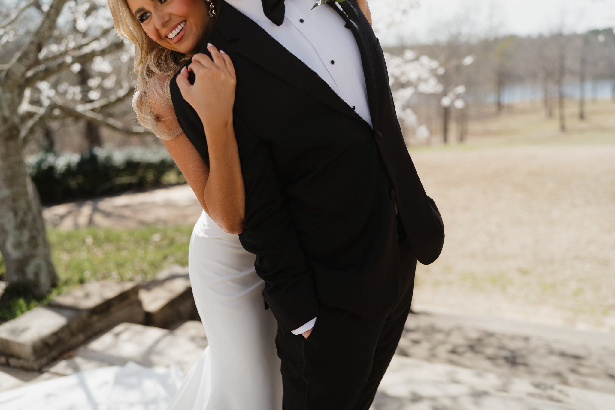 A bride and groom in wedding attire sharing an intimate moment outdoors, with the bride wearing a white dress and the groom in a black tuxedo, against a backdrop of trees and natural scenery in Memphis, TN. 