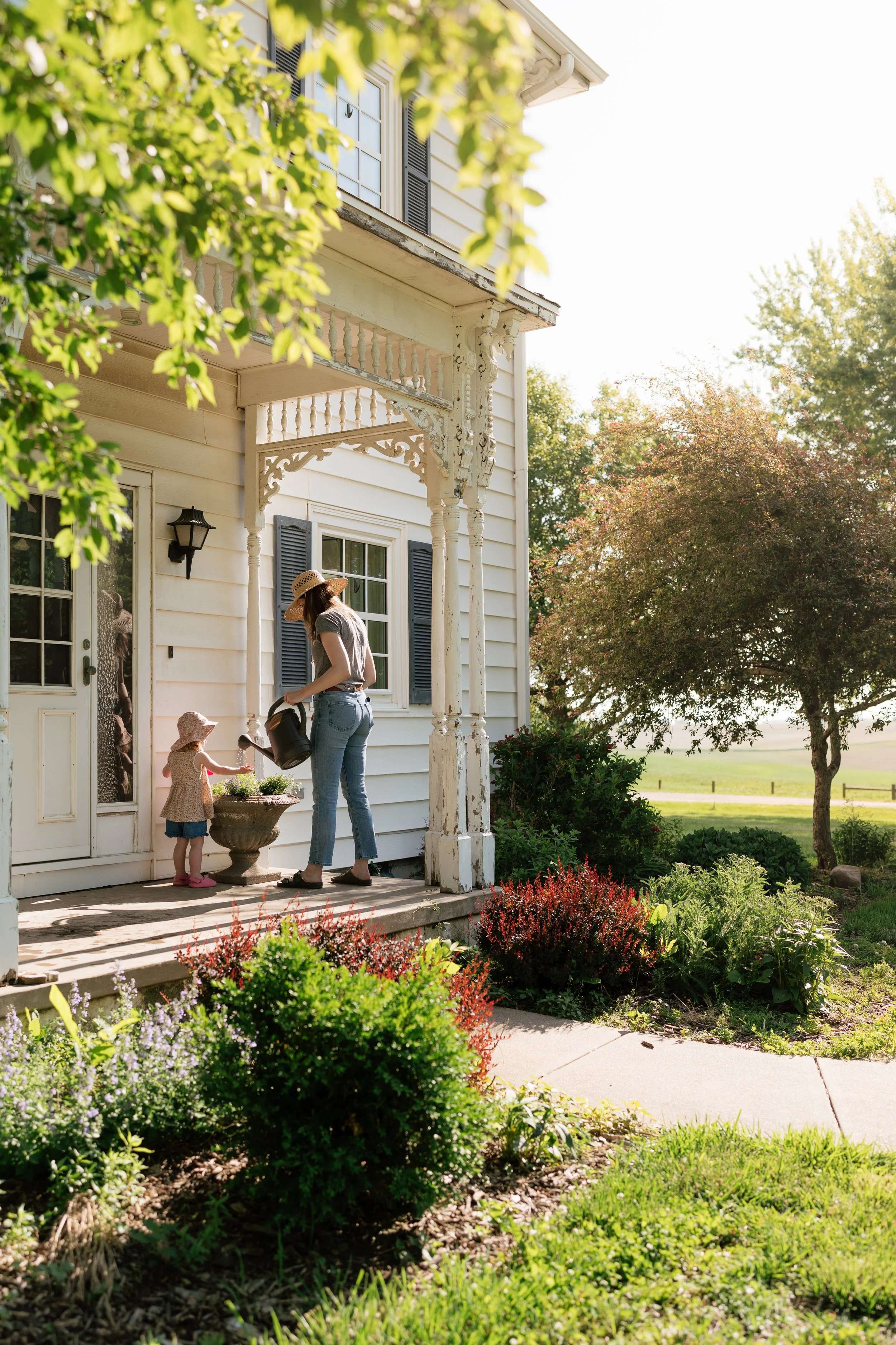 A woman and two young girls are on the porch of a white house with black shutters, surrounded by greenery. The woman is watering plants while the girls look on. The scene is sunny with trees in the background on a farm in Tennessee. 