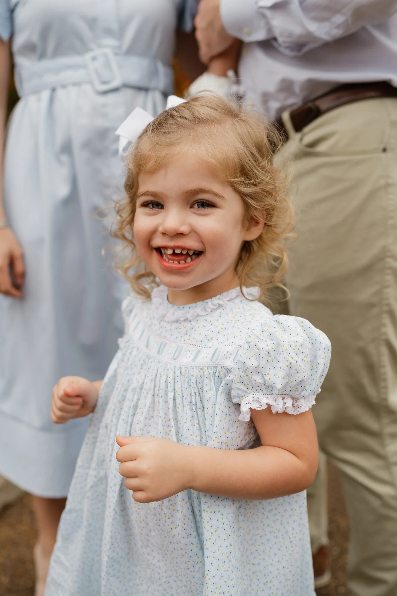 A young girl with curly blonde hair and a white bow, smiling and showing missing front teeth, wearing a white dress with subtle colorful dots, standing outdoors among adults in Memphis, TN. 