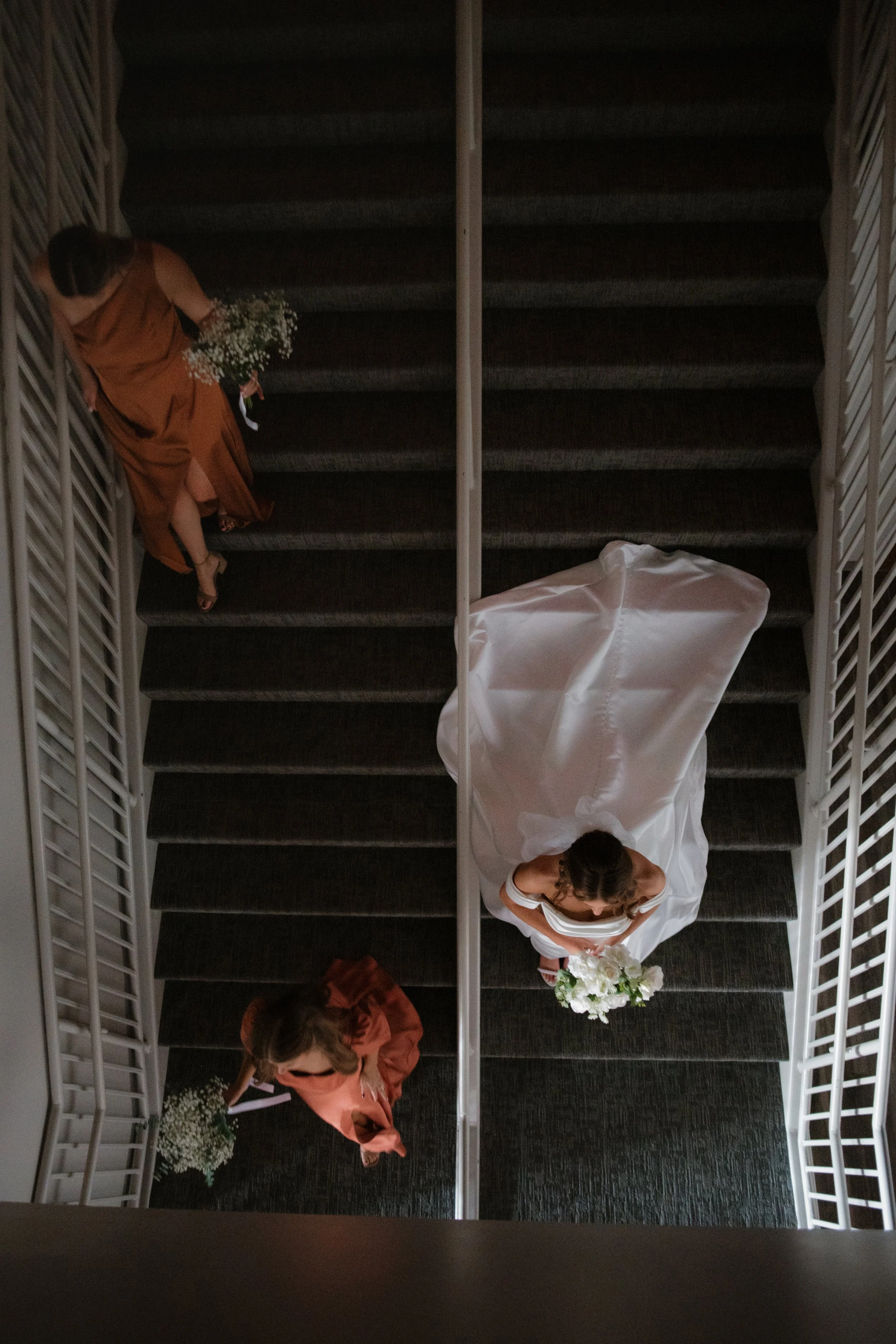 A bride in a white wedding dress holding a bouquet of white flowers walking up a staircase, with several women accompanying her, some in coral dresses, on a dark carpeted staircase viewed from above in Memphis, TN. 