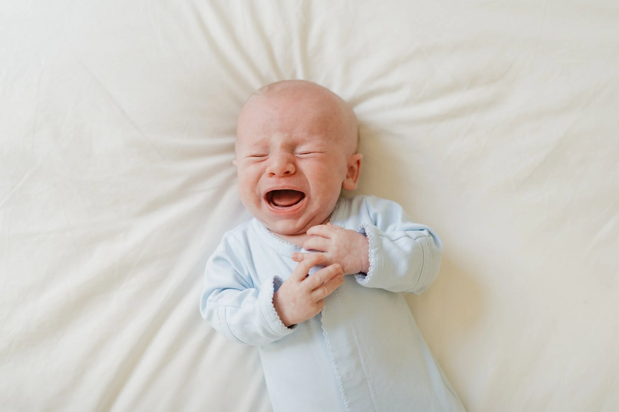 A crying newborn with a bald head, lying on a white sheet and wearing a long-sleeved white shirt with gray accents in Memphis, TN. 