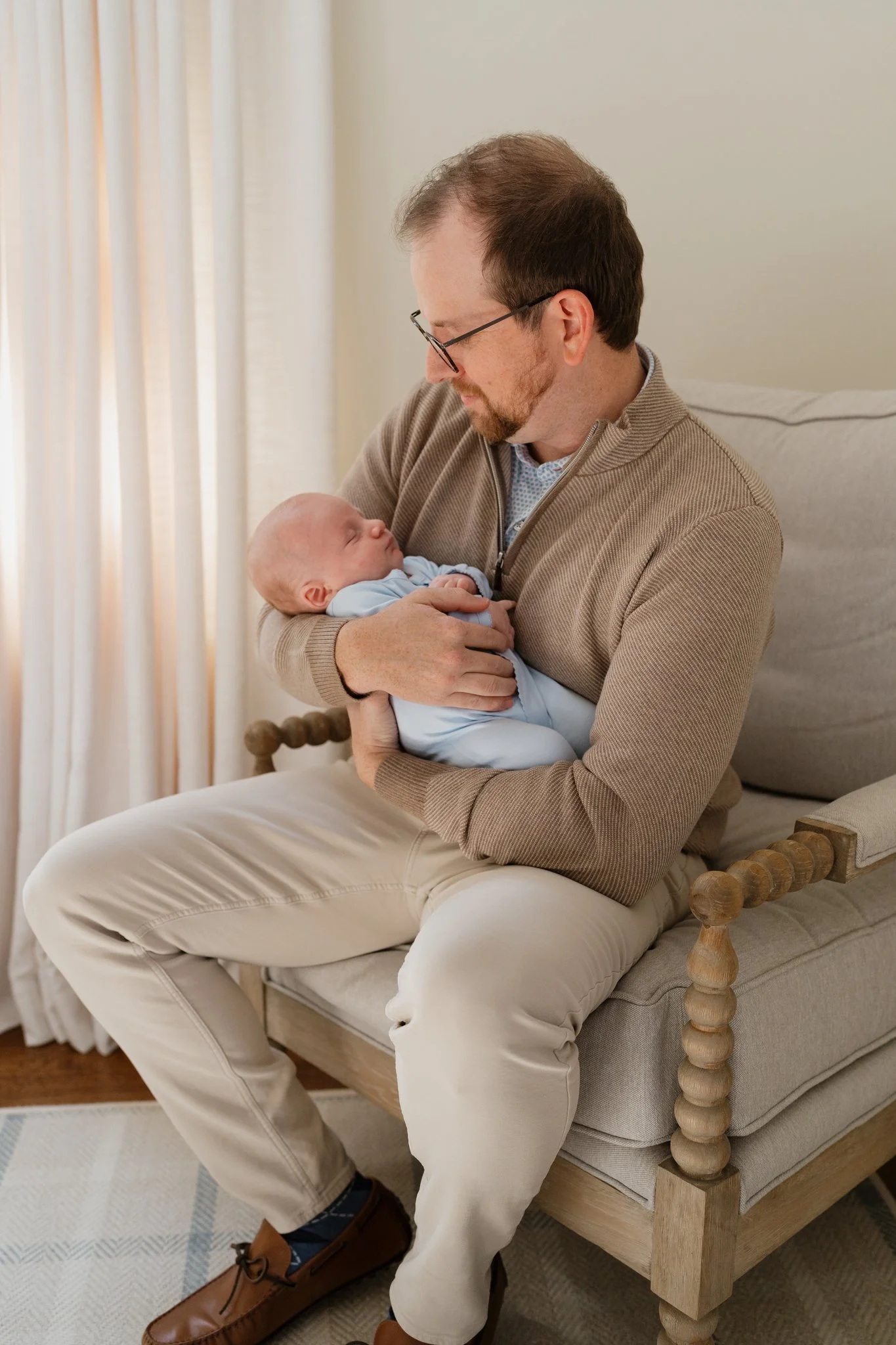 A man with glasses holding a sleeping baby in his arms while sitting on a chair in a room with cream walls and curtains in Memphis, TN. 