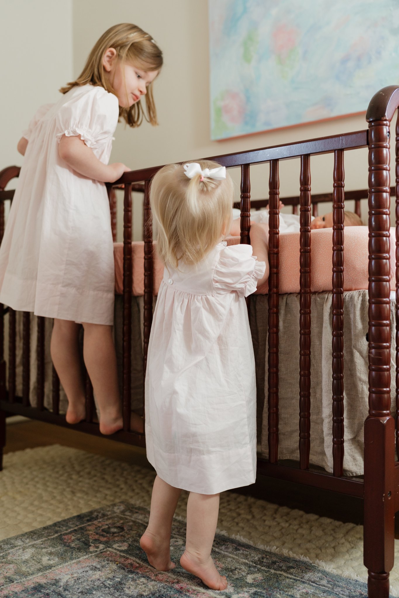 Two young girls, one with light brown hair and the other with blonde hair, are standing by a wooden crib. The girl with brown hair is leaning on the crib, while the blonde girl, wearing a white dress and a white bow in her hair, is looking inside the