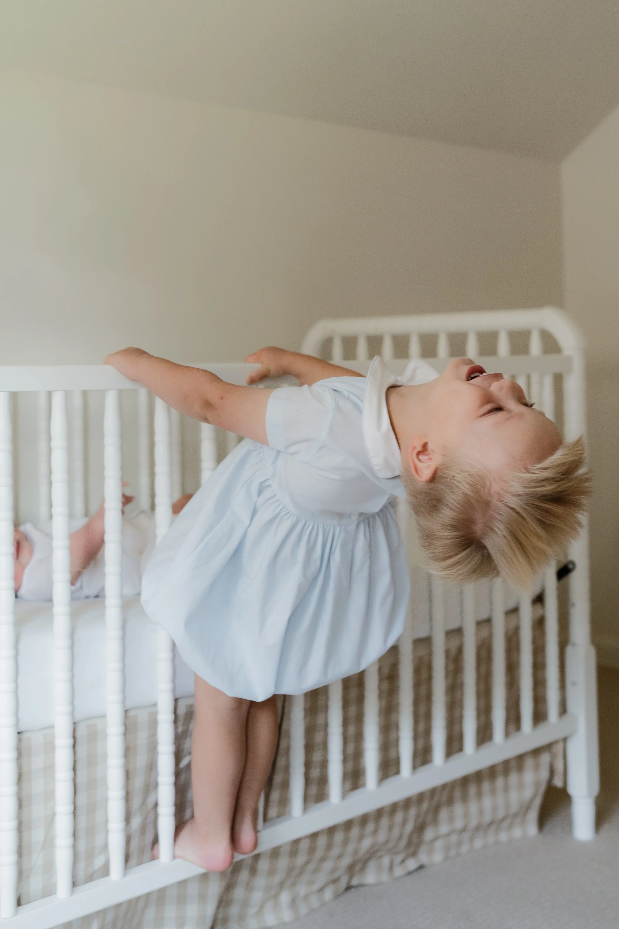 A young child with blonde hair wearing smock, standing in a crib, smiling and reaching out with one arm in Memphis, TN. 