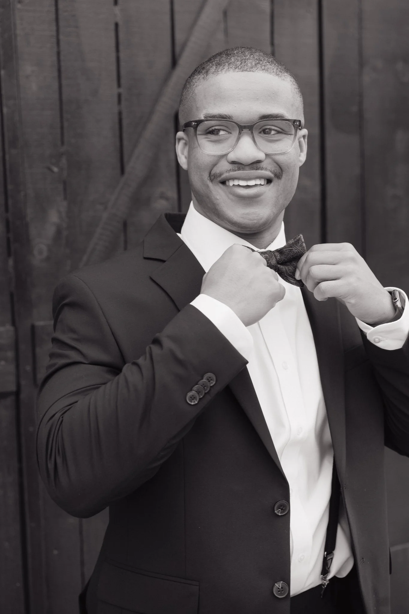 A smiling man adjusting his bow tie while wearing glasses, a suit, and a watch, standing against a wooden background in Memphis, TN. 