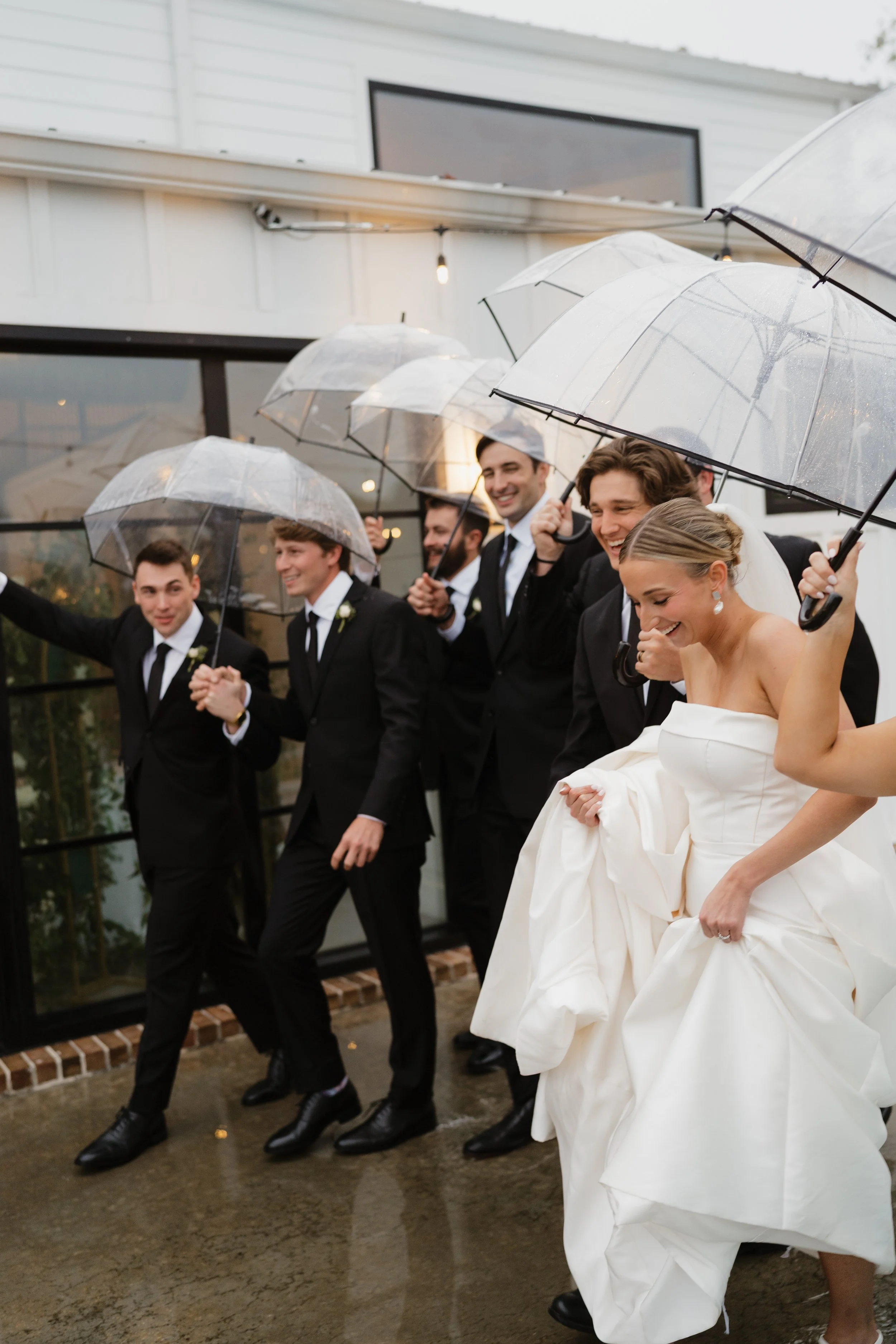 A group of wedding guests, mostly men in suits, holding transparent umbrellas and smiling as they walk outside in the rain, with a woman in a wedding gown also smiling in Memphis, TN. 