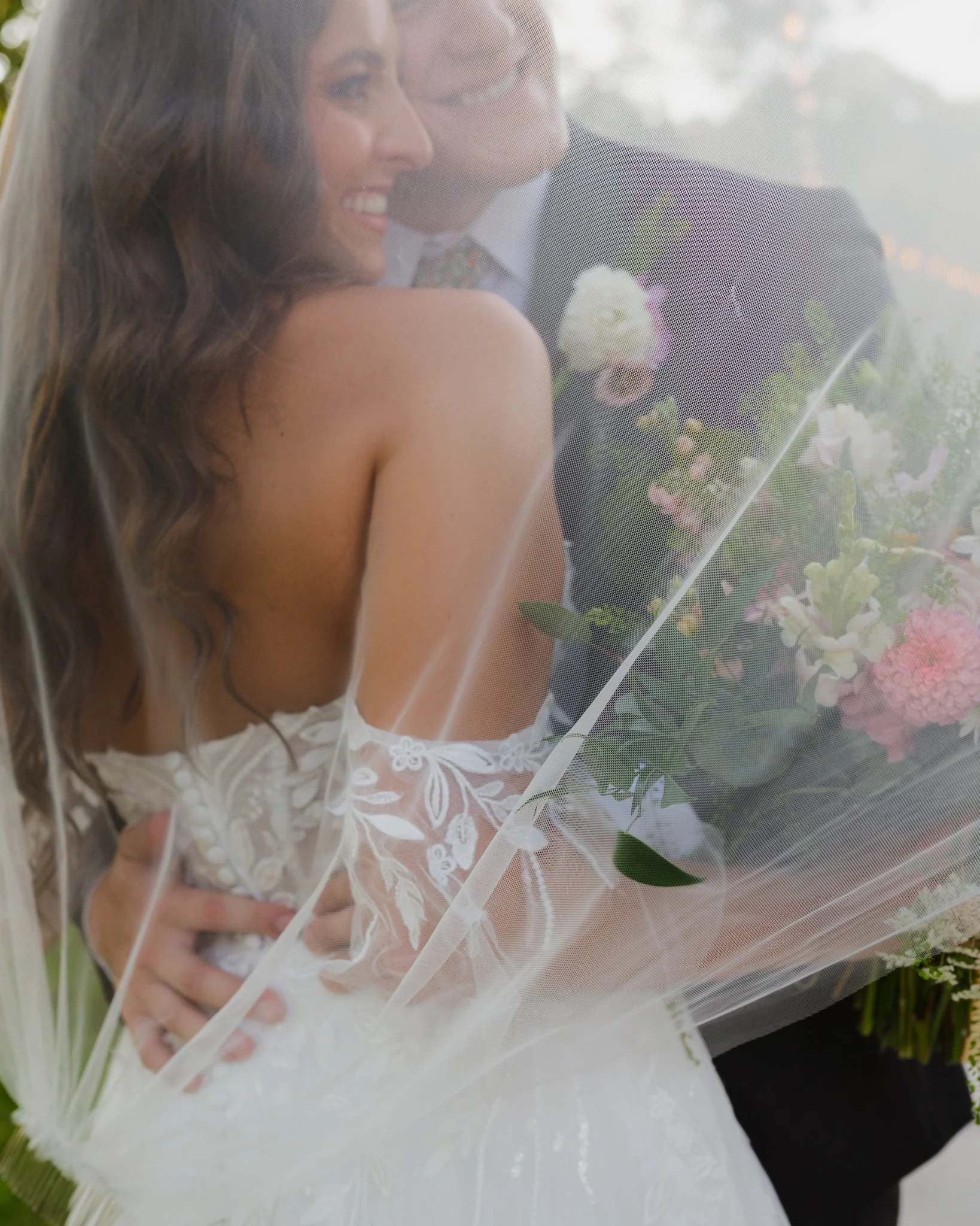 A bride with long wavy brown hair and a groom in a dark suit, smiling behind a sheer veil, holding a bouquet of white and pink flowers outdoors in Memphis, TN. 
