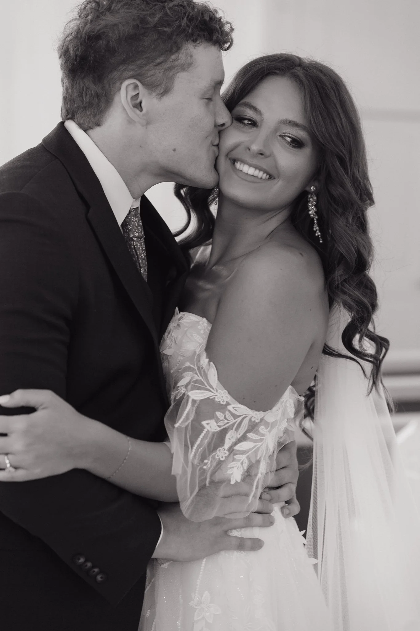 A black and white photo of a couple, with the man kissing the woman's cheek. The woman is smiling and wearing a lace wedding dress with floral details and earrings. The man is wearing a suit and tie, with his arm around the woman in Memphis, TN. 