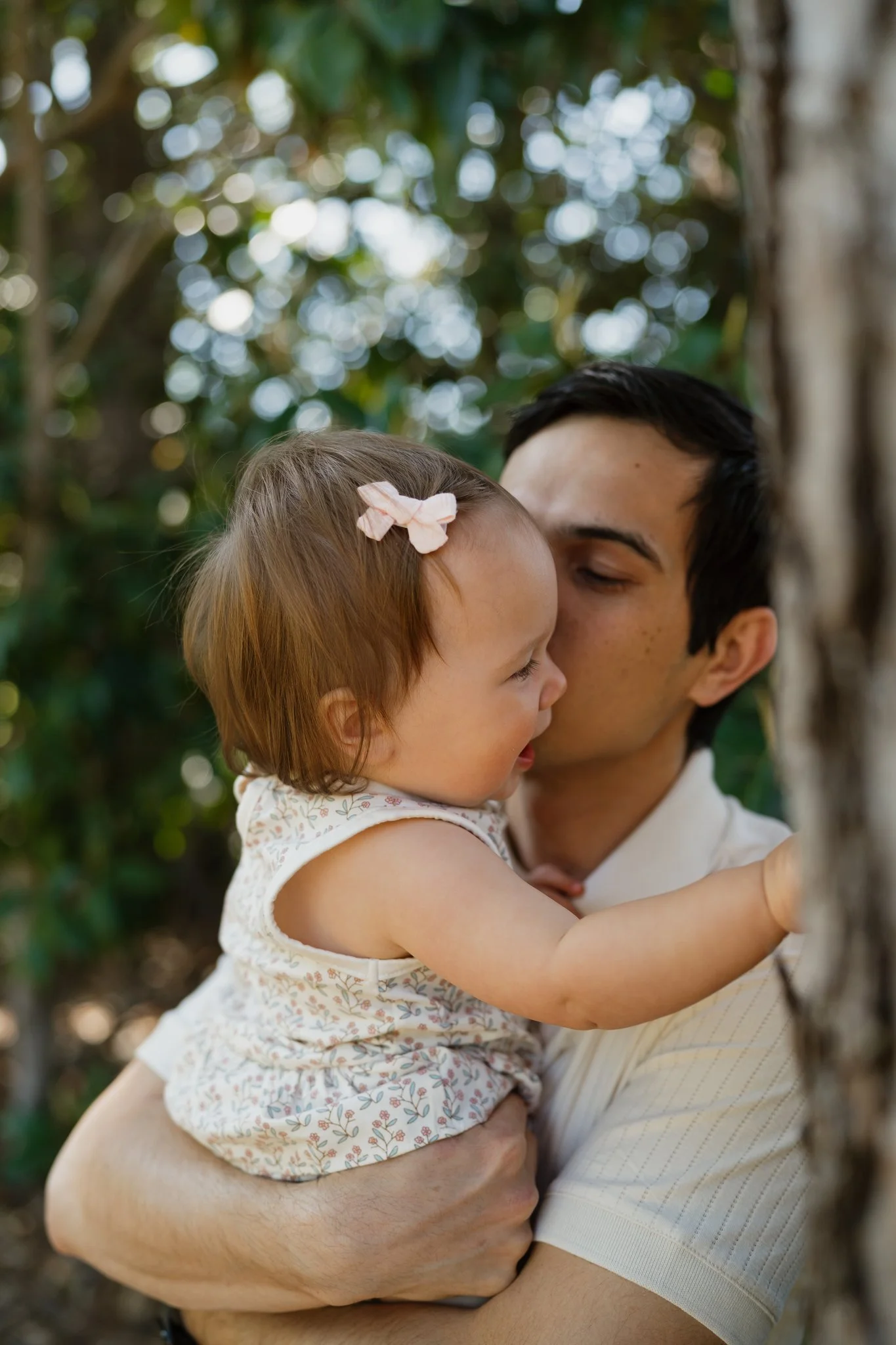 A man holding a young girl outdoors with green trees and sunlight in the background.