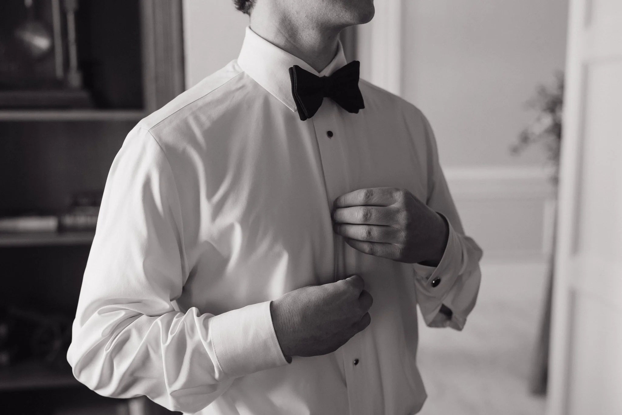 A man adjusting his tuxedo shirt, wearing a black bow tie, in a formal setting with shelves and decor in the background in Memphis, TN. 