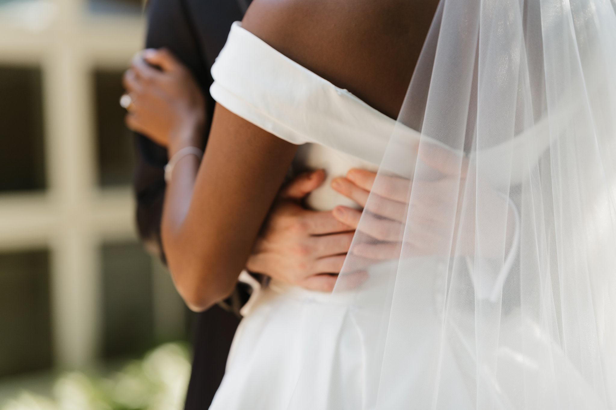 Close-up of two people, one with dark skin and one with light skin, holding each other at a wedding. The person in white is likely the bride, wearing a veil, and the person in dark clothing is possibly the groom. They are embracing, with the bride's 
