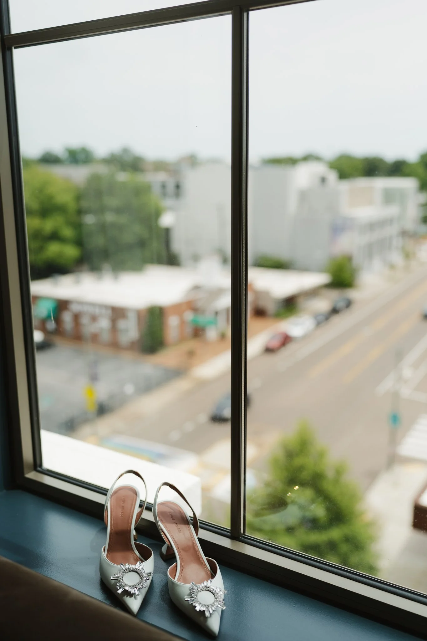 Pair of white wedding shoes with decorative rhinestone embellishment on a windowsill, with a blurred urban street view outside in Memphis, TN. 
