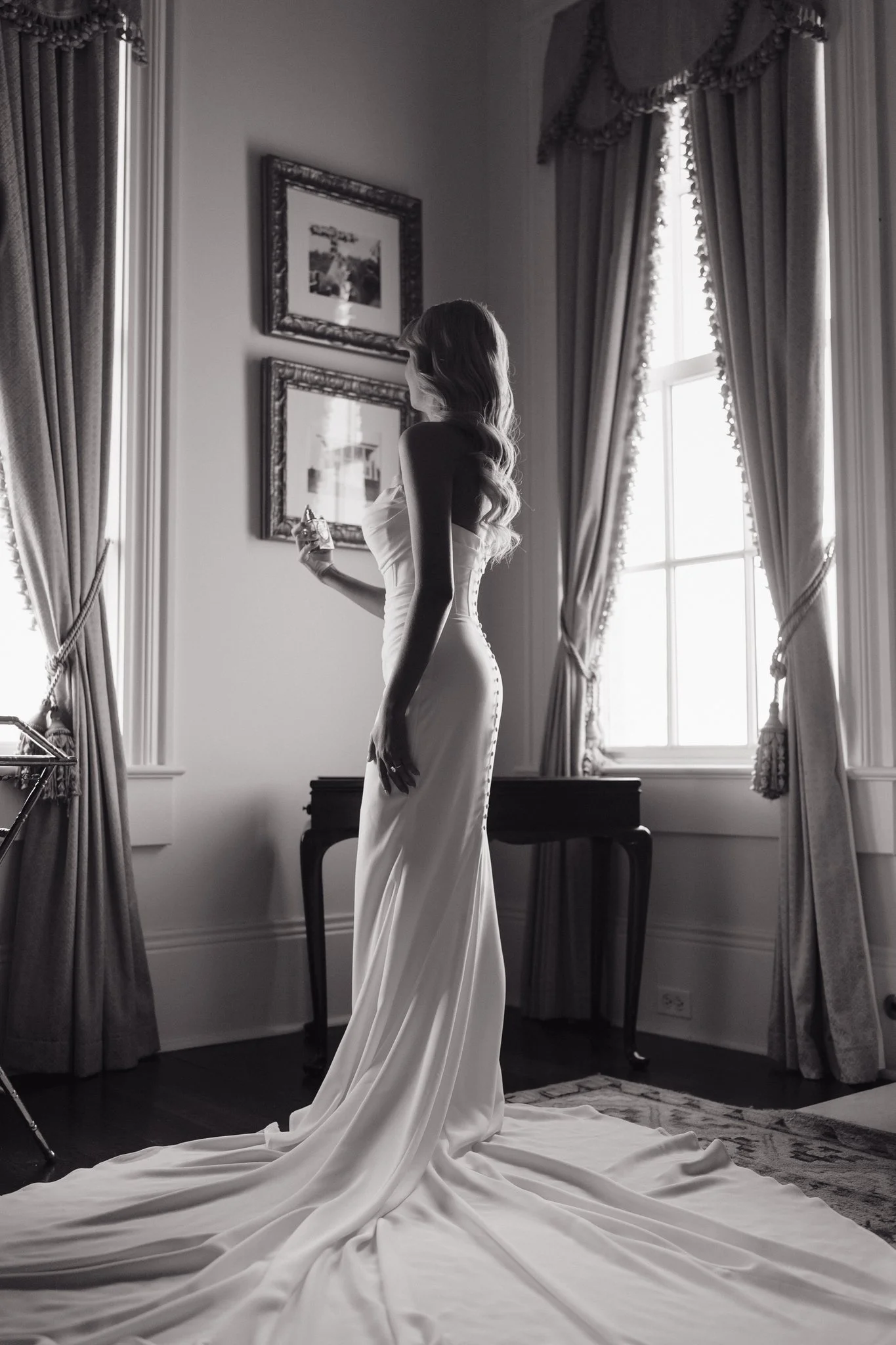 Woman in a wedding dress holding a perfume bottle in a well-lit room with large windows and curtains, framed pictures on the wall, and a rug on the floor, in Memphis, TN. 