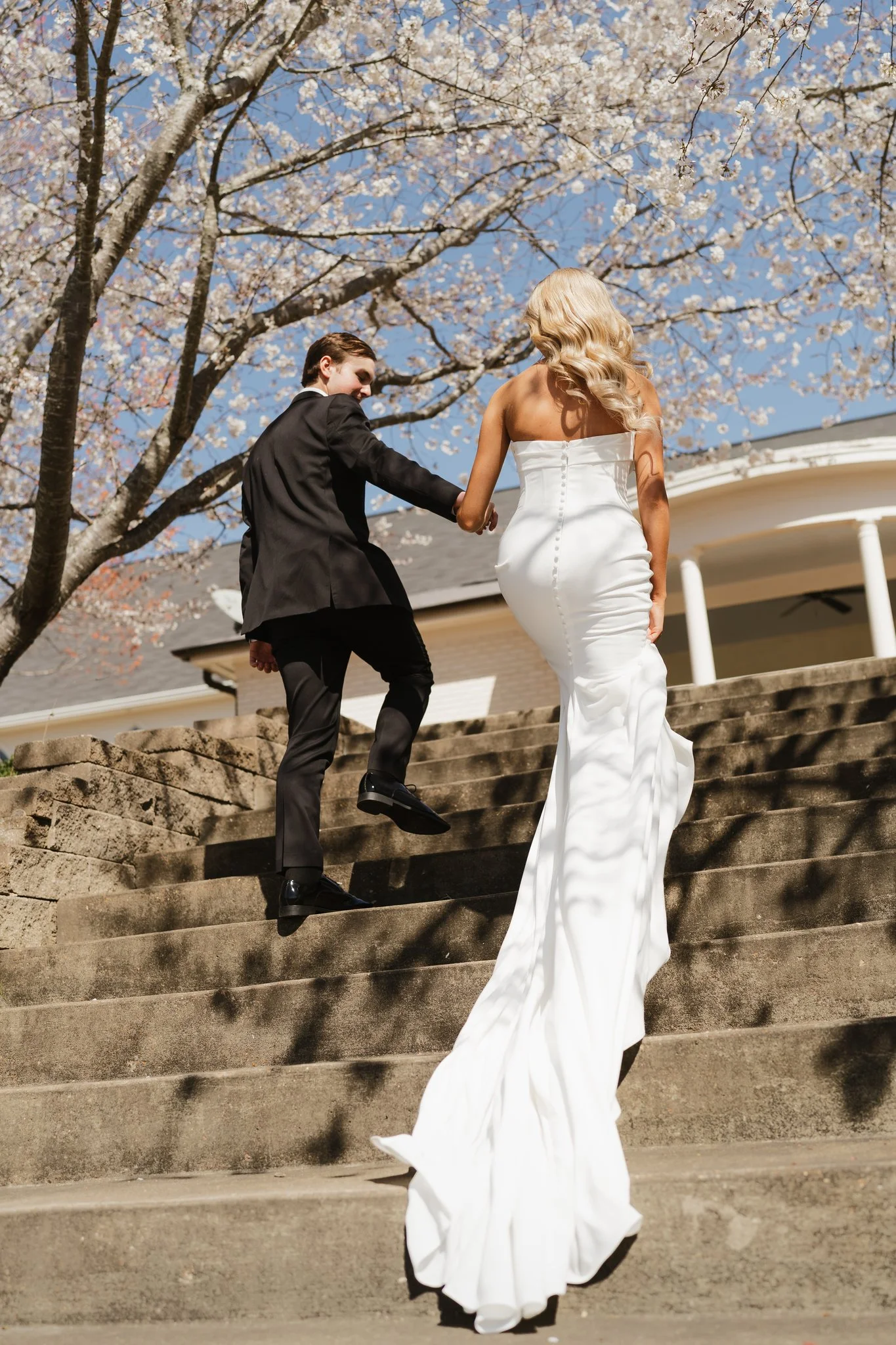 A bride and groom walking up stone steps outdoors under a blooming cherry blossom tree, with the bride wearing a white strapless wedding gown and the groom in a black suit, holding hands in Memphis, TN. 
