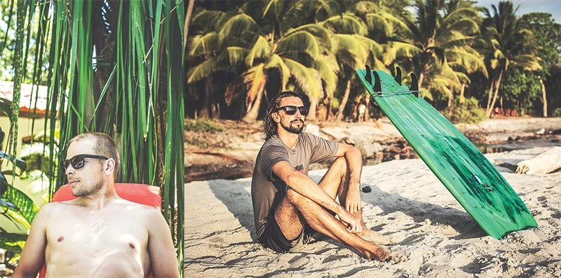 Two men relaxing on a tropical beach, one lying on a lounge chair in front of green plants, and the other sitting on the sand with a surfboard propped up beside him, palm trees in the background.