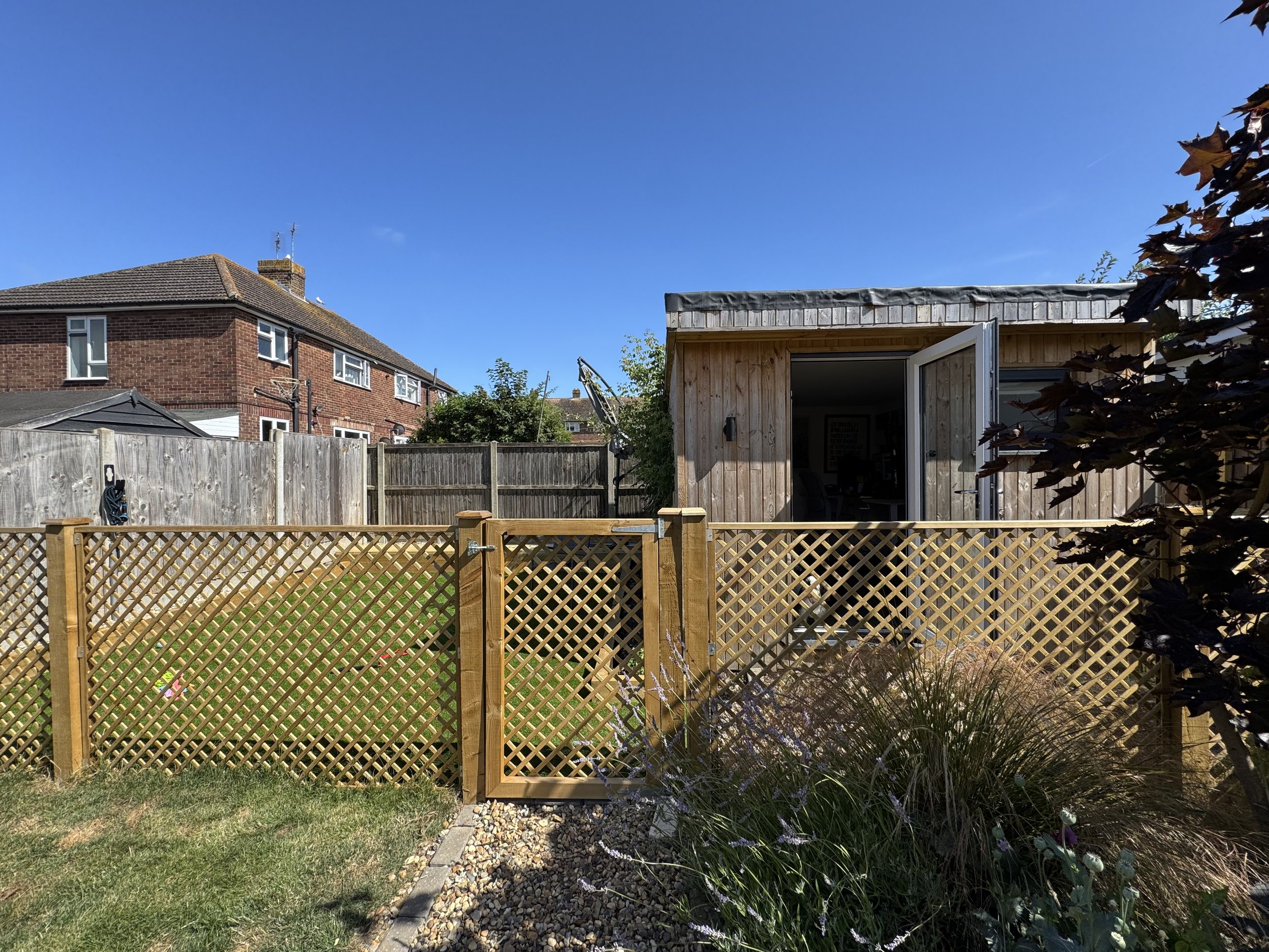 A small wooden garden shed with a glass door, situated behind a wooden lattice fence in a backyard with a lawn, plants, and a paved path. A neighboring brick house is visible in the background under a clear blue sky.