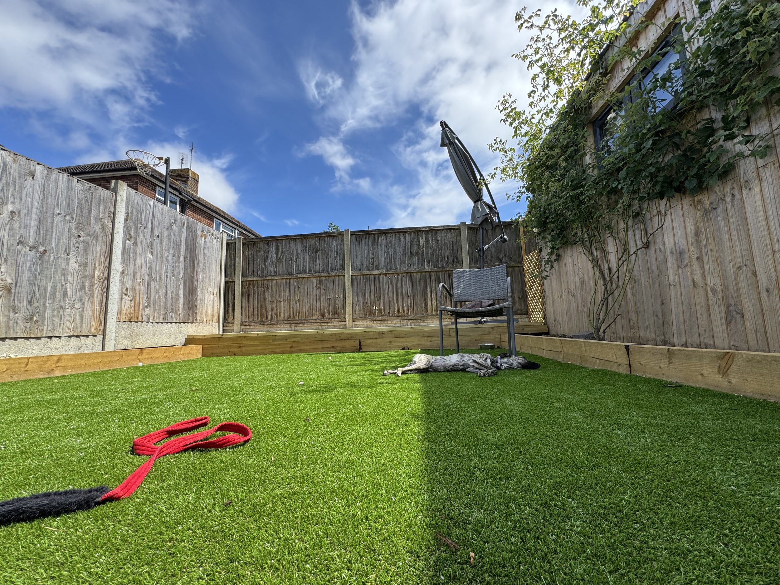 A backyard with freshly laid green artificial grass, a red leash, a dog laying on the ground, a black chair, a closed patio umbrella, and a wooden fence under a partly cloudy sky.