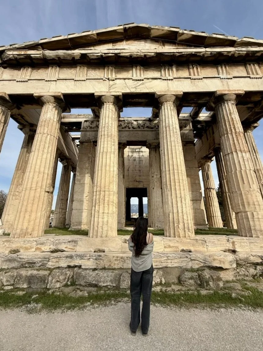 A woman with long dark hair, wearing a gray top and black pants, stands in front of the Parthenon, an ancient Greek temple with tall marble columns, on a clear day.