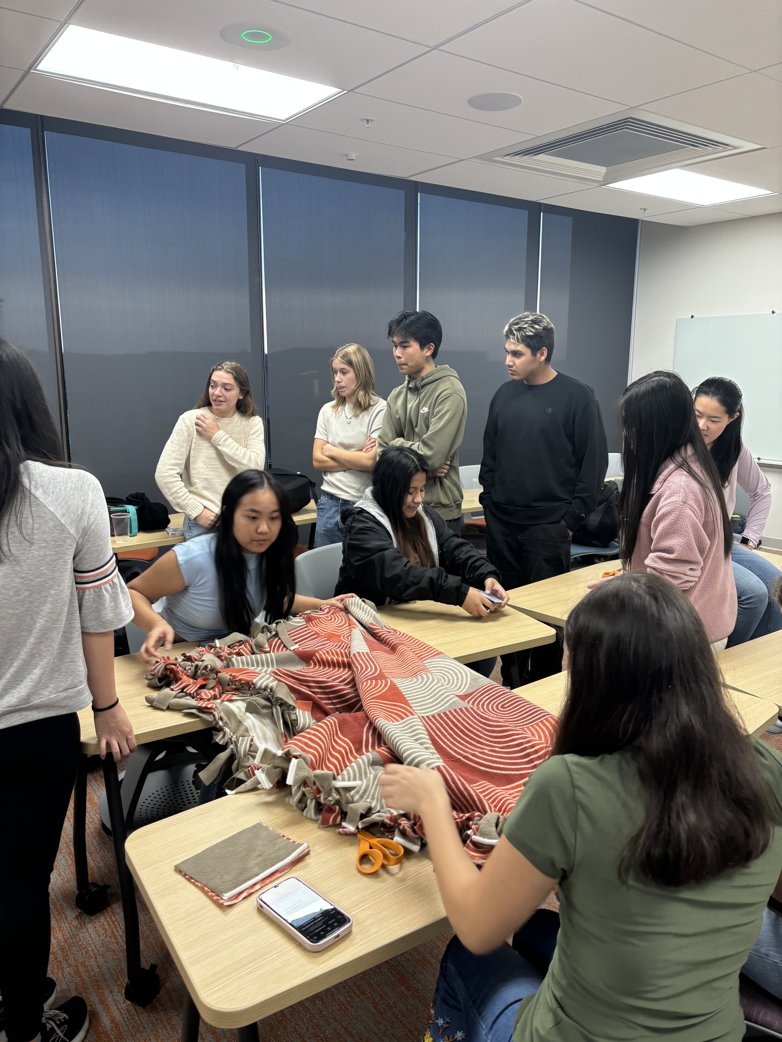 Group of students gathered around tables with fabric, scissors, and a smartphone, working on a craft or project in a classroom.