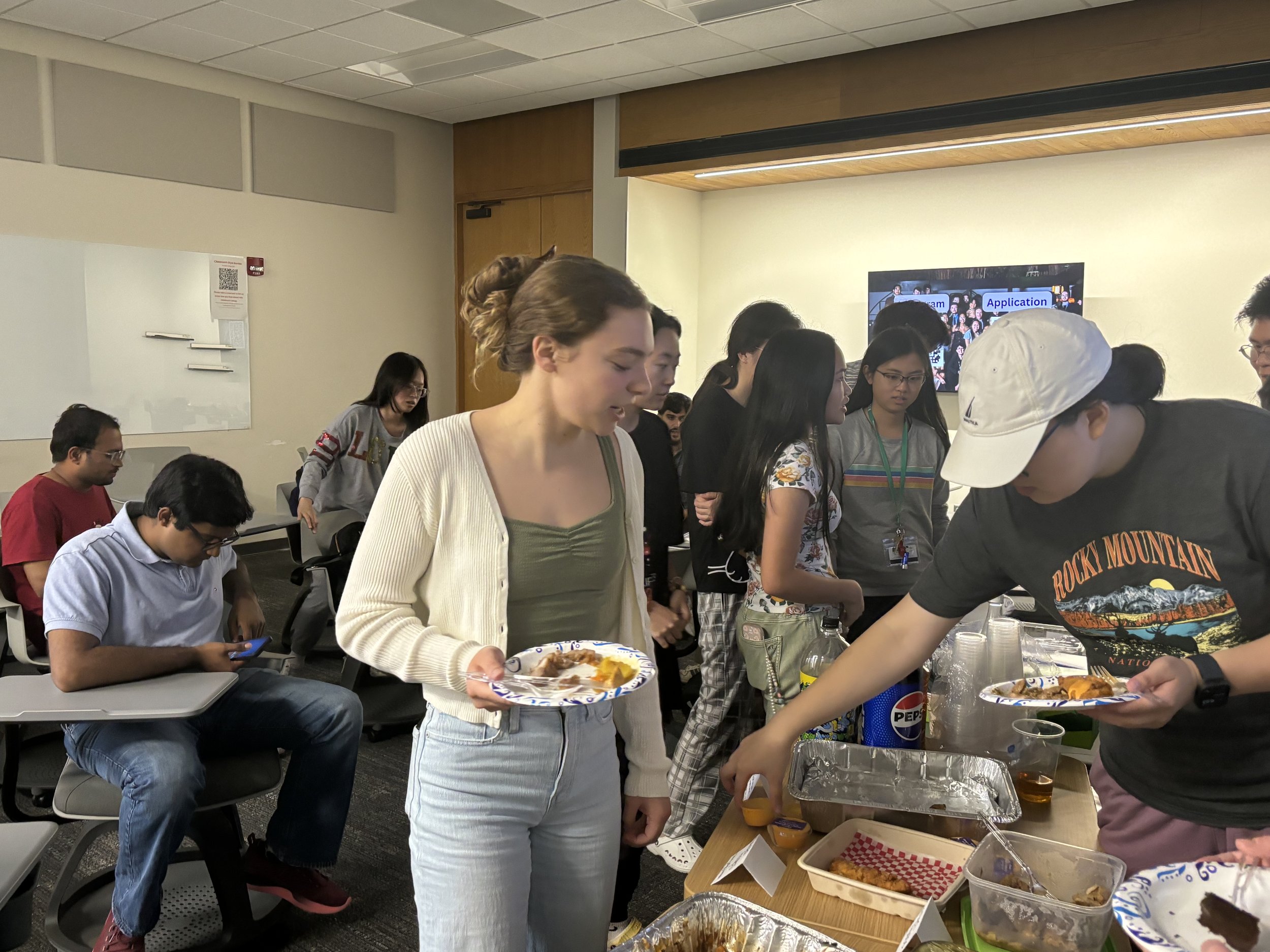 Group of people in a classroom, some seated and some standing, getting food from a buffet table.
