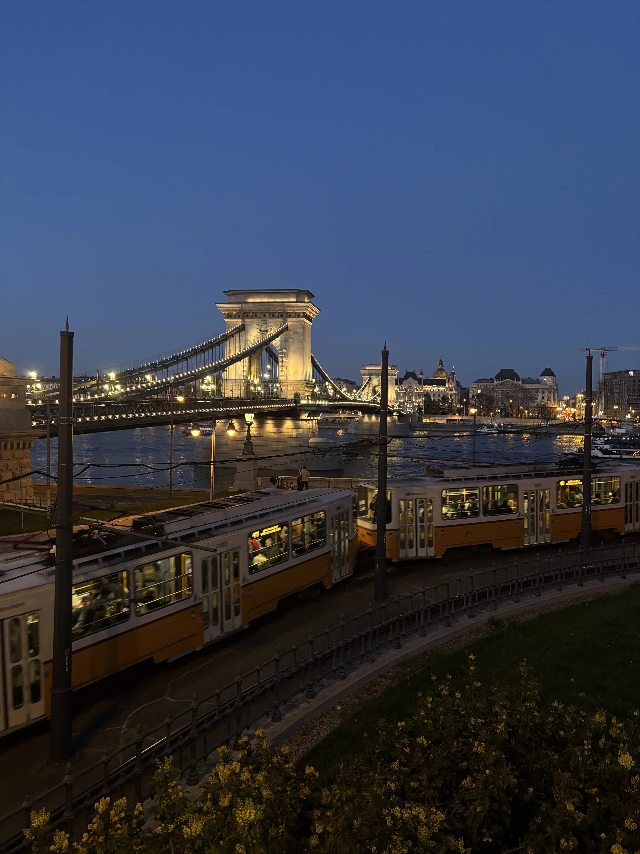 Nighttime view of Chain Bridge over the Danube River in Budapest, Hungary, with illuminated buildings and two yellow and white trams passing by in the foreground.