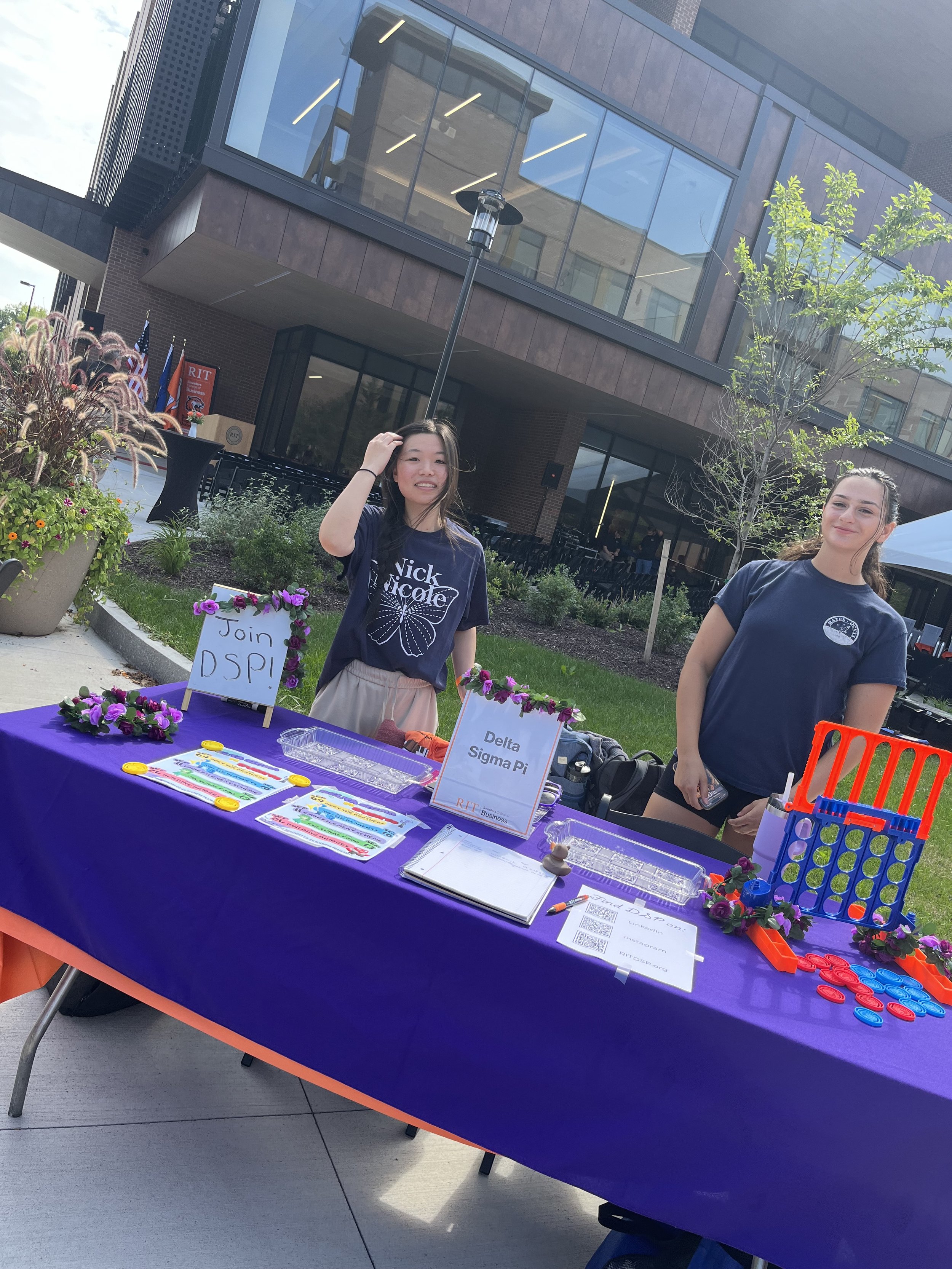 Two young women standing behind an outdoor table with purple tablecloth, decorated with flowers and signs that read "Join DSP!" and "Delta Sigma Pi." The table has papers, plastic discs, and a Connect Four game. One woman is wearing a dark blue T-shi