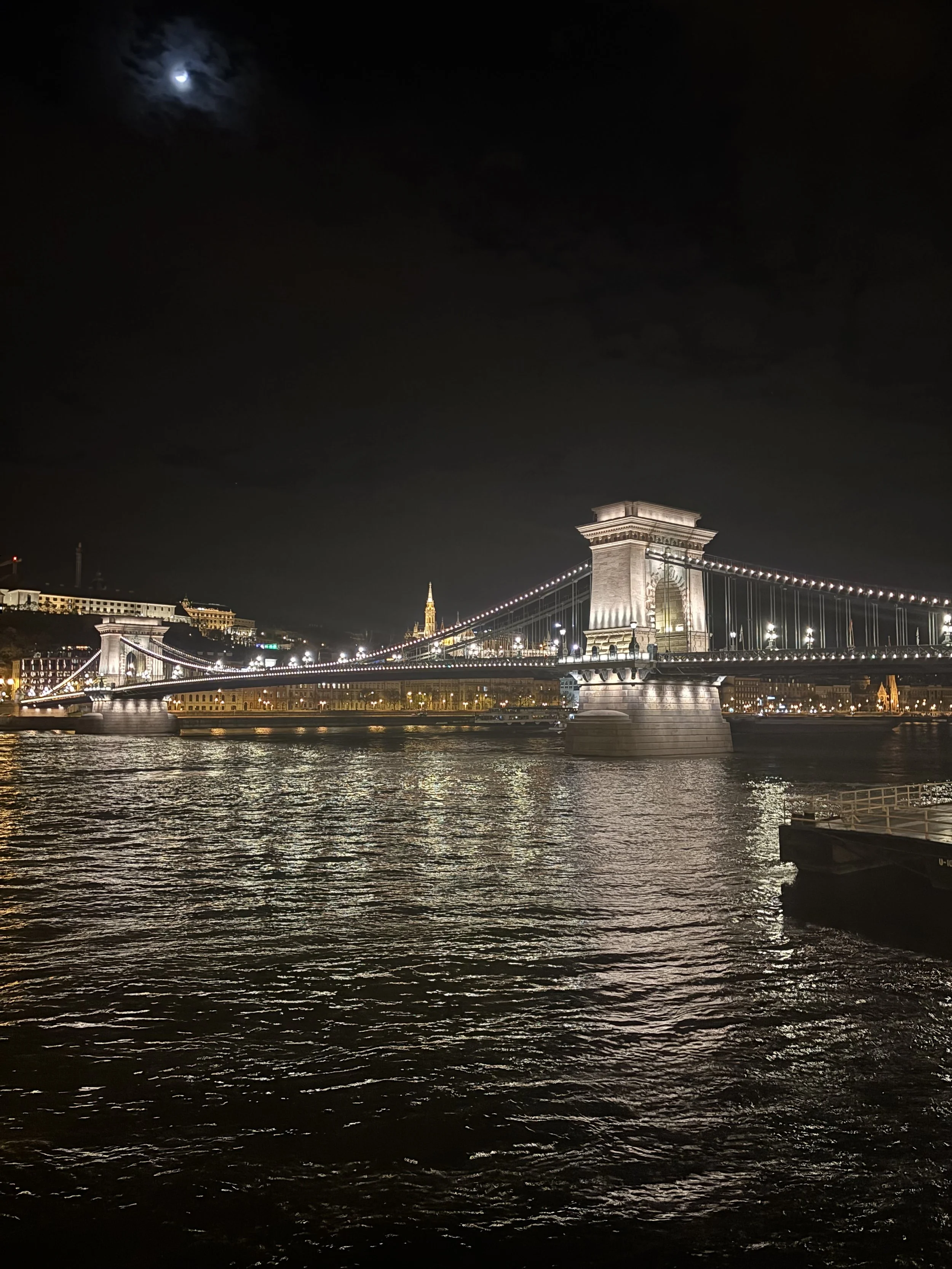 Night view of the Brooklyn Bridge illuminated with city lights in the background and a half moon in the cloudy sky reflected on the water.