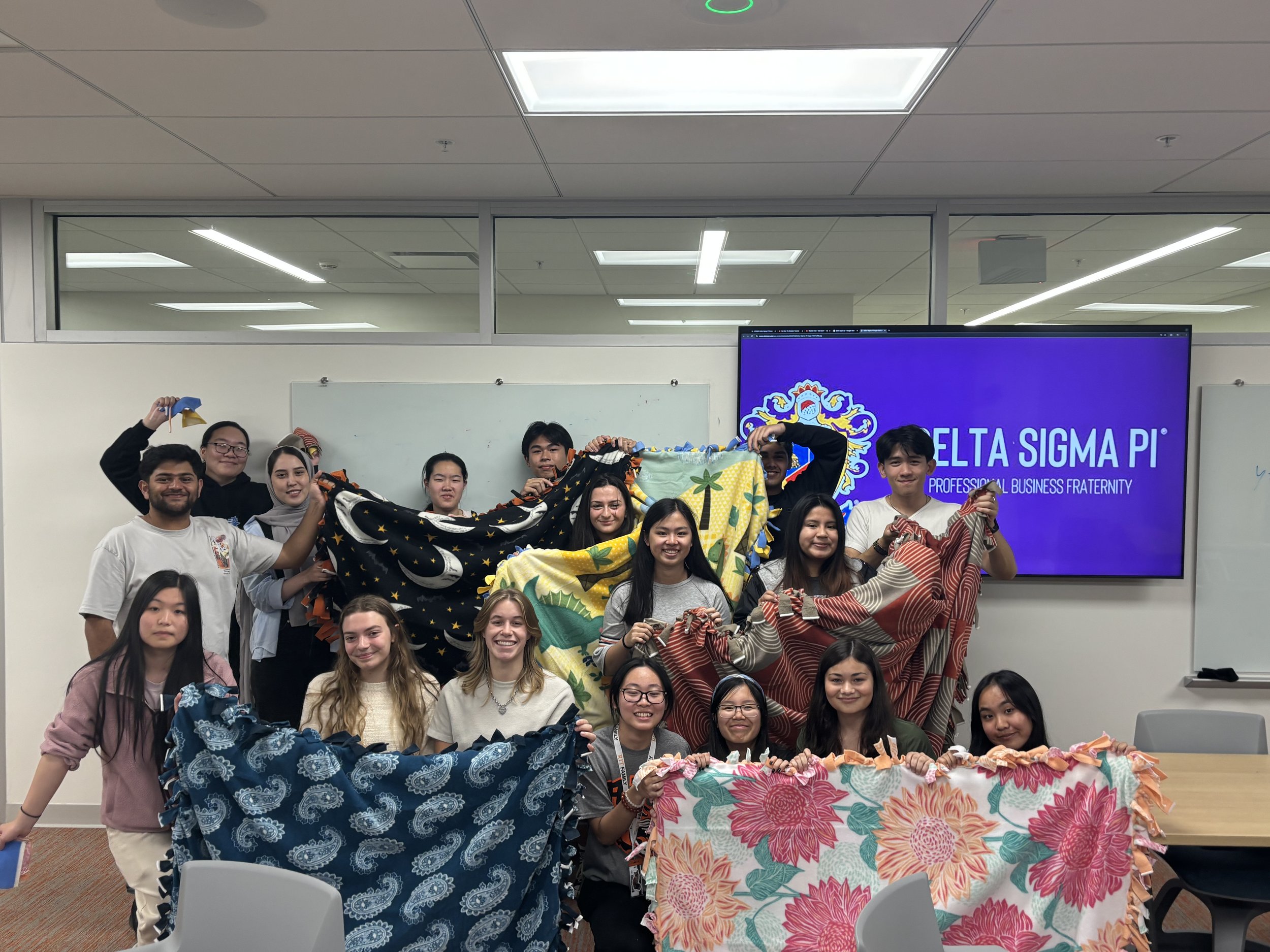 Group of young adults holding colorful patterned fabric, standing in a classroom in front of a screen displaying 'Delta Sigma Pi' and a whiteboard.