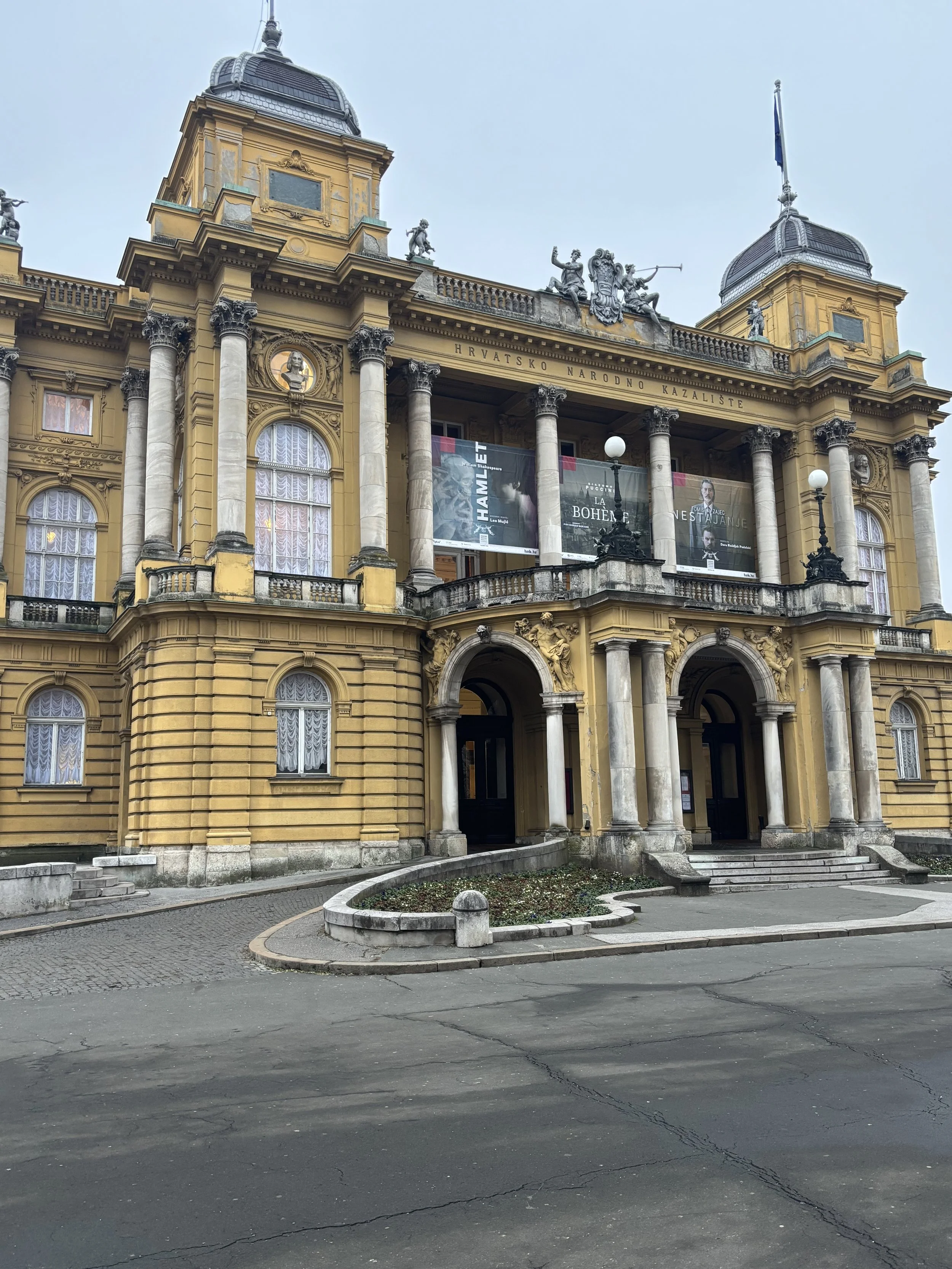 Historic yellow building with ornate architecture, columns, statues, and banners, with a Croatian flag on top, under a cloudy sky.