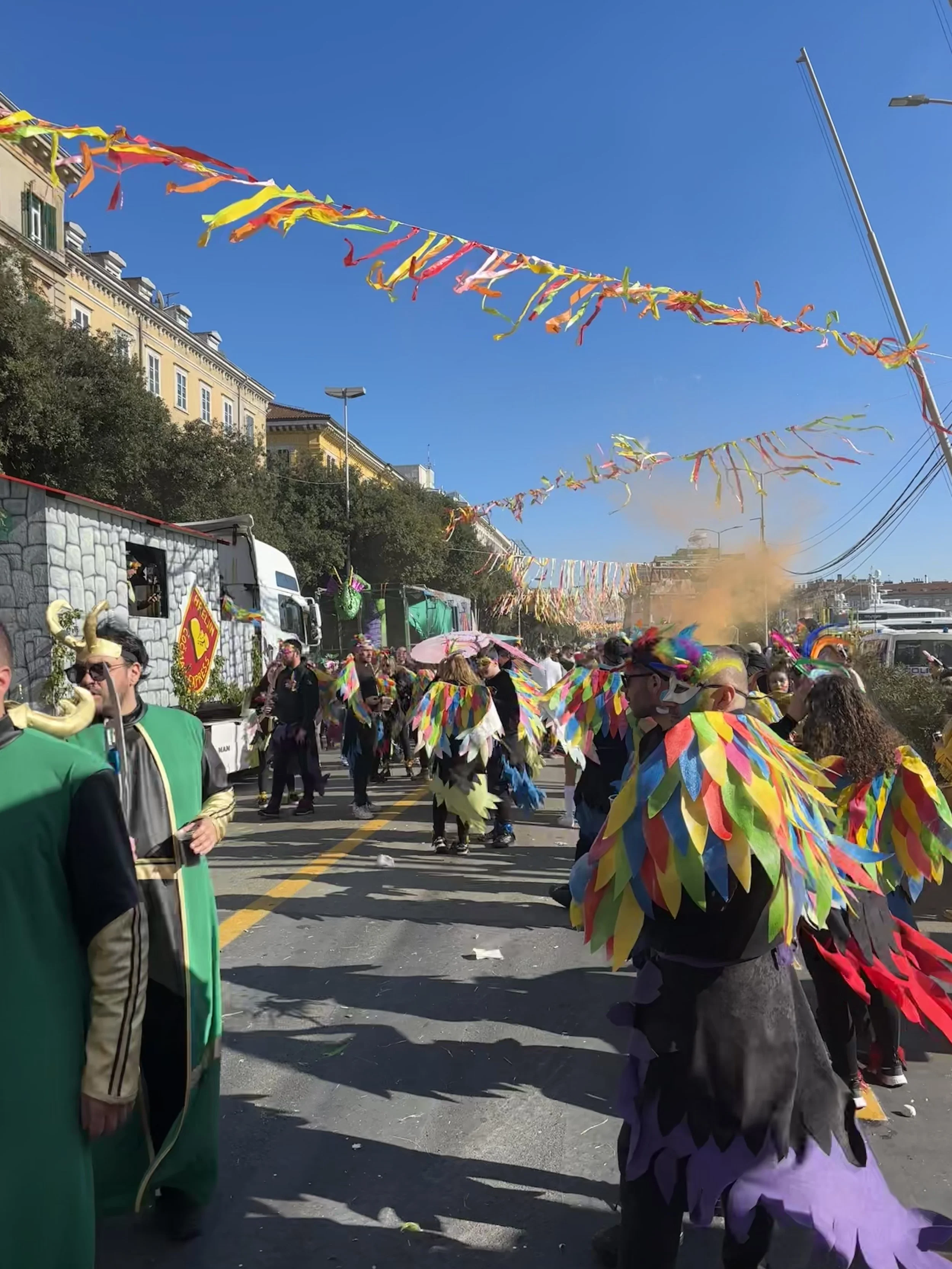 People dressed in colorful costumes with feathered capes participating in a street parade during a festival with bright blue sky and colorful streamers overhead.
