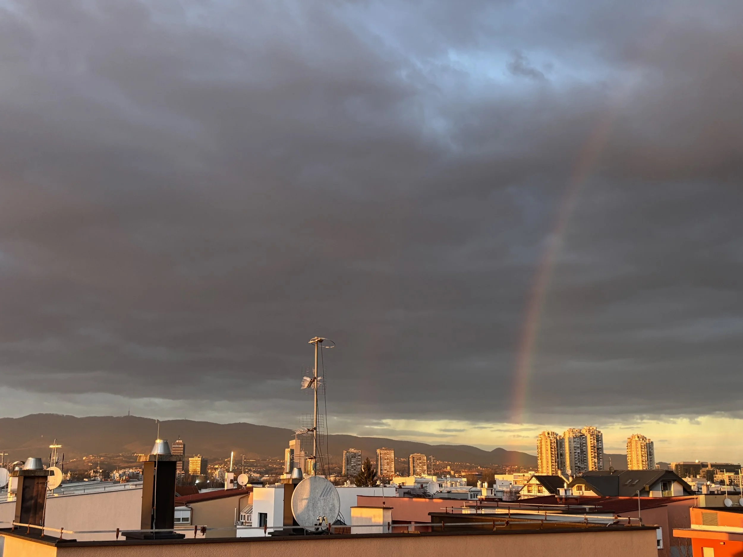 Cityscape with rooftops, tall buildings, and a rainbow in a cloudy sky at sunset.