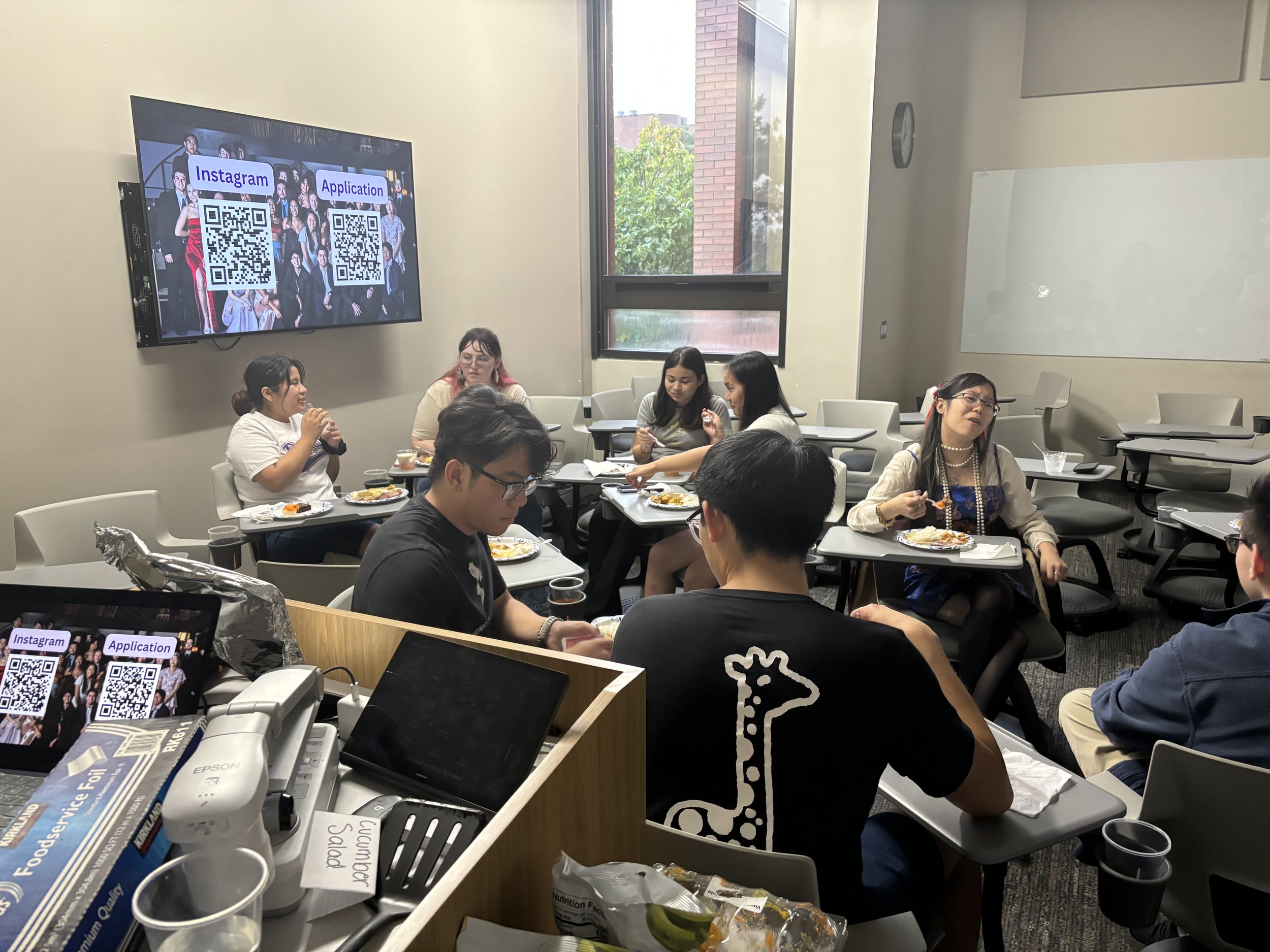 Group of young adults eating and talking in a classroom conference room with a large window, whiteboard, and a digital screen displaying QR codes for Instagram and application.