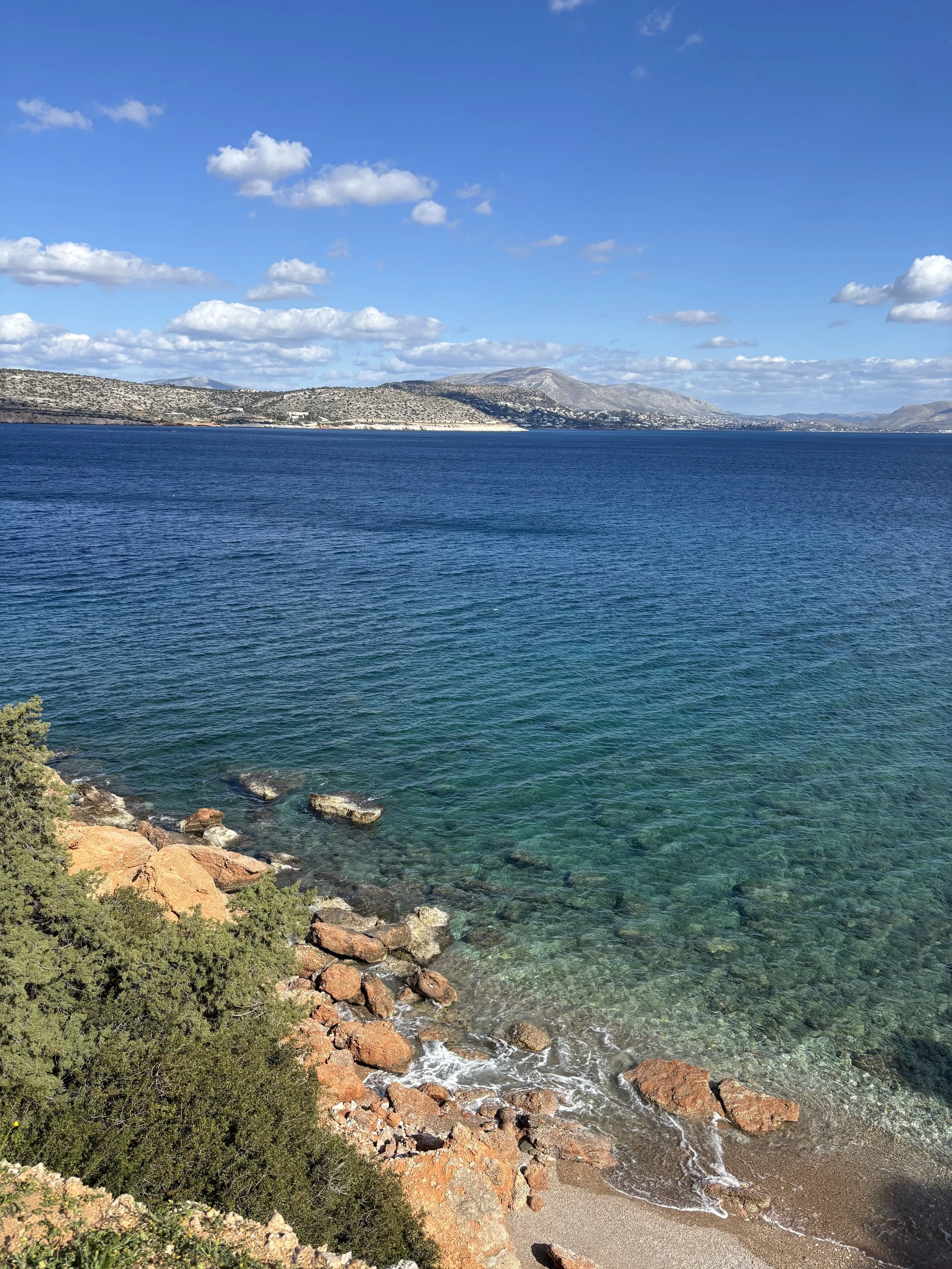 Scenic view of a rocky beach with clear blue water, green shrubbery, and distant hills under a partly cloudy sky.