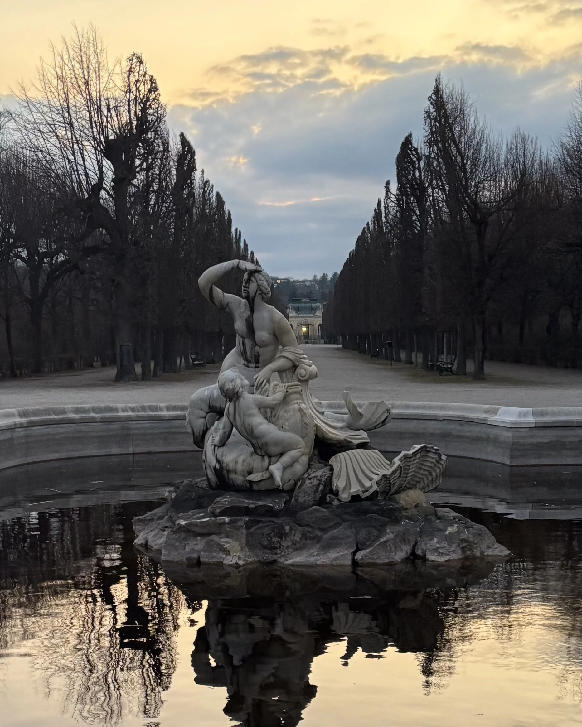 Sculpture of mythological figures in a fountain with trees and a pathway in the background at sunset.
