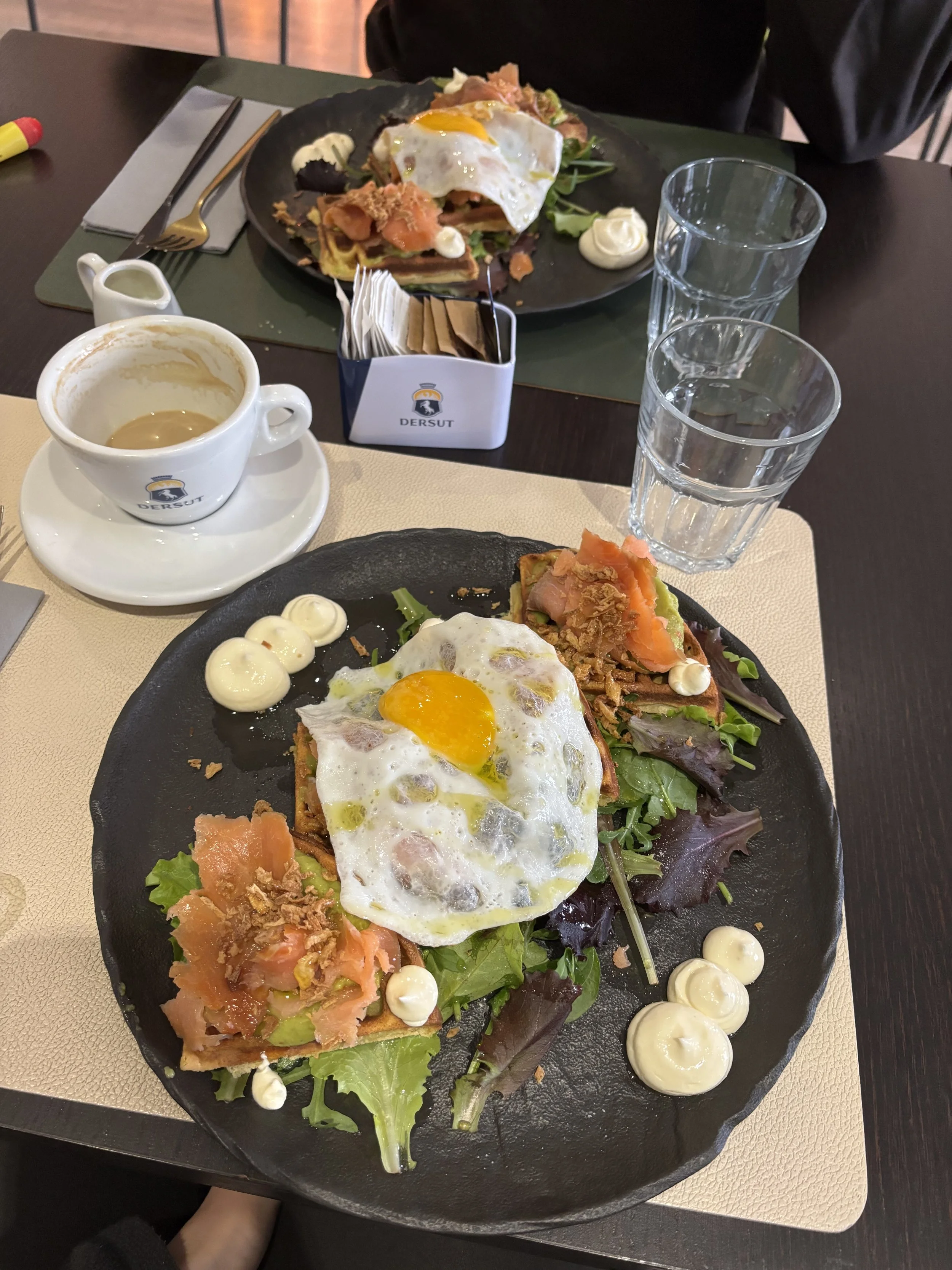 Plate of salad topped with a fried egg, with dollops of mayonnaise or dressing, on a black plate. In the background, a second similar plate is visible. There is a glass of water, a cup with coffee or espresso, and a small container of condiments on a