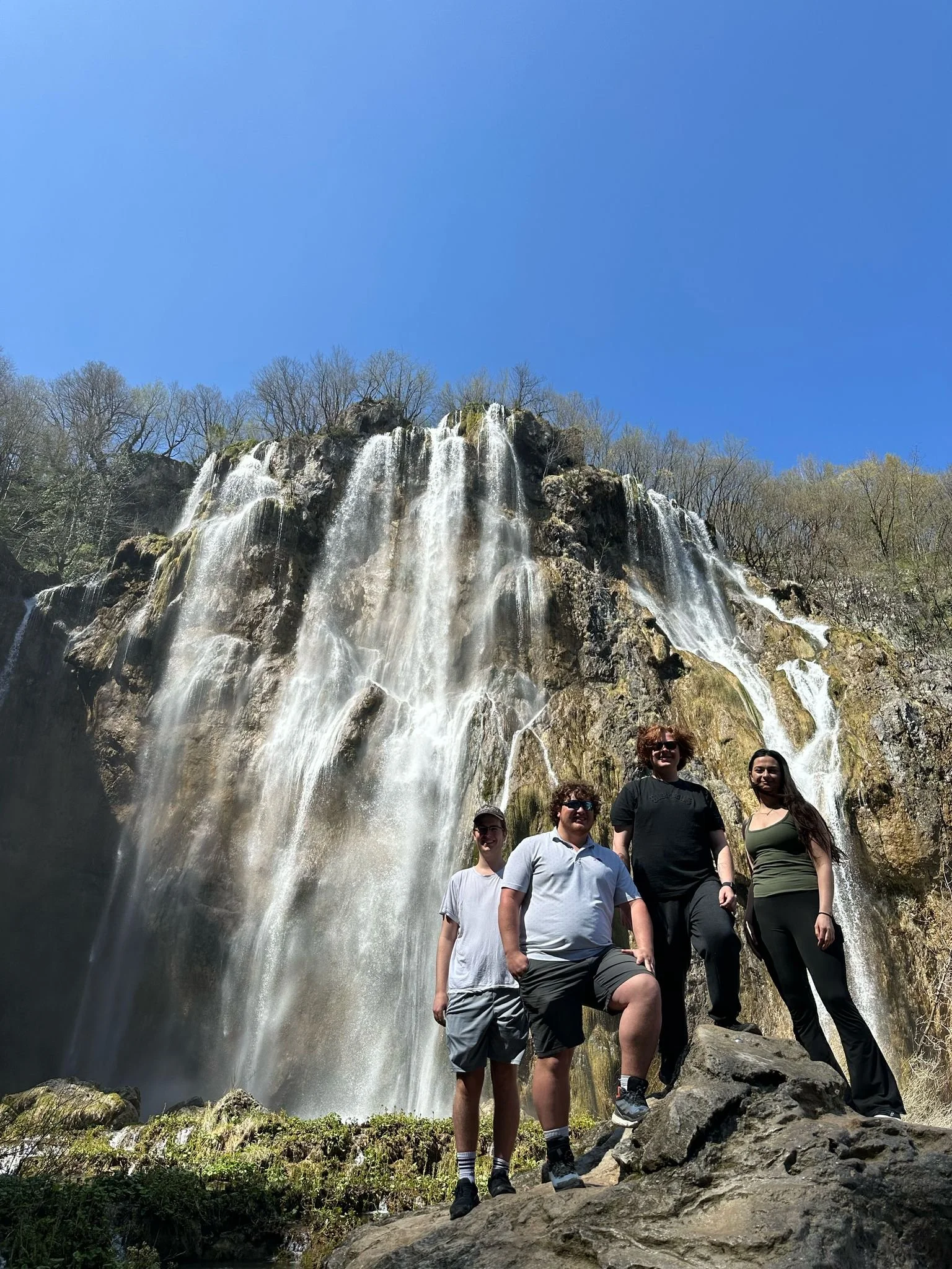 Four people standing on rocks in front of a waterfall during daytime with a clear blue sky and trees in the background.