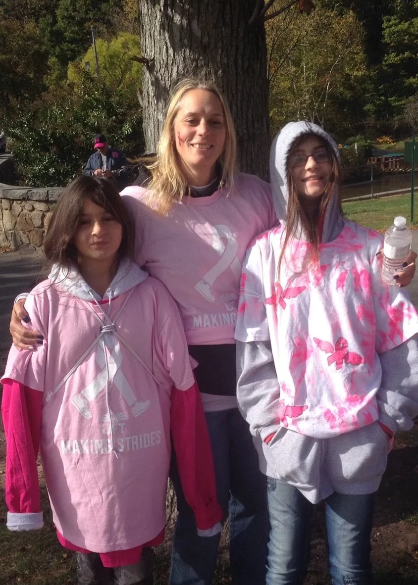 Three girls and a woman standing outdoors near a large tree, wearing pink themed T-shirts and sweatshirts, with autumn leaves in the background.