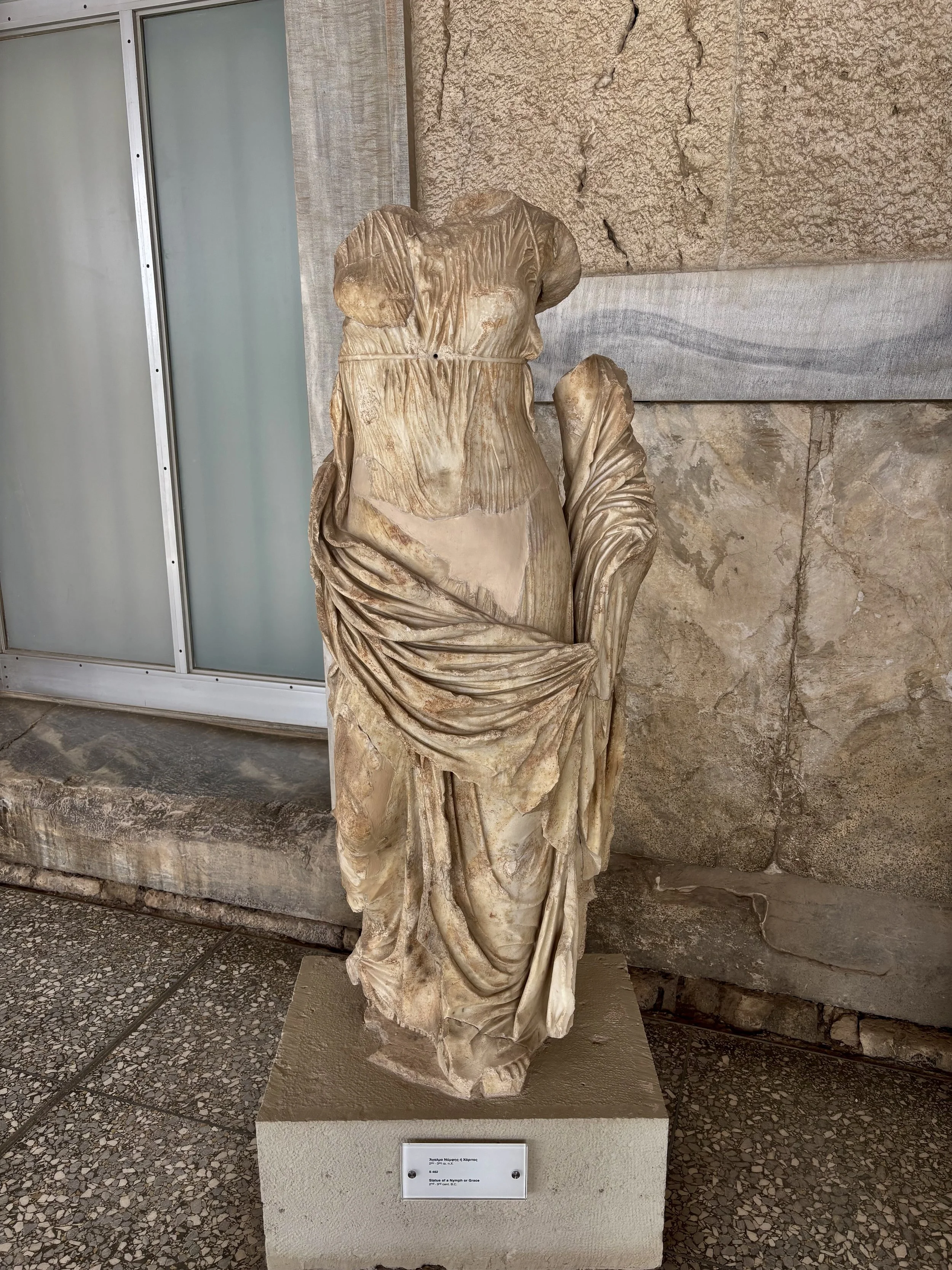 Ancient marble sculpture of a draped female figure, missing head and arms, displayed on a beige pedestal.