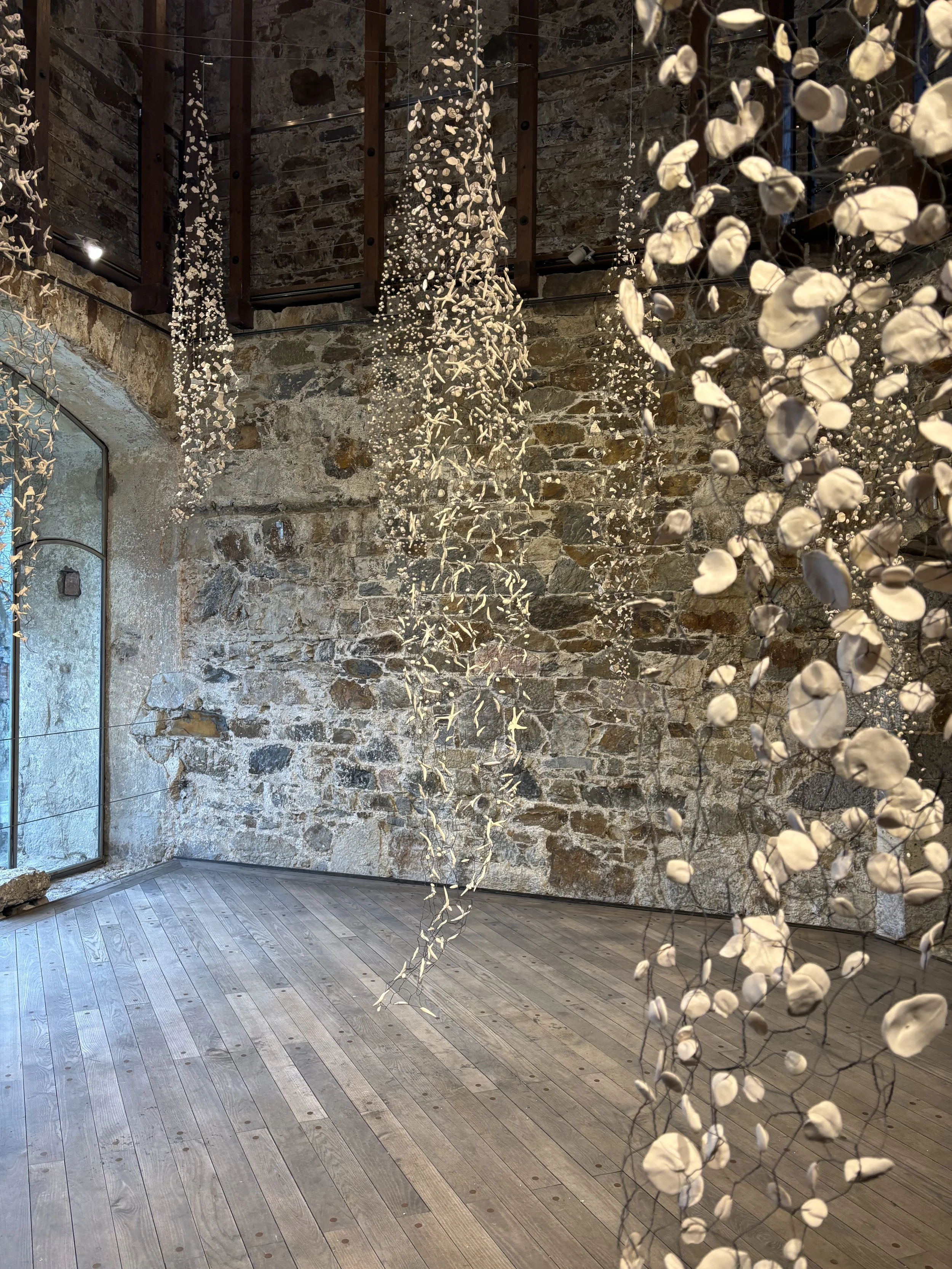 Interior of a rustic stone-walled room decorated with hanging white floral art installations, with wooden flooring and a large industrial-style window to the left.