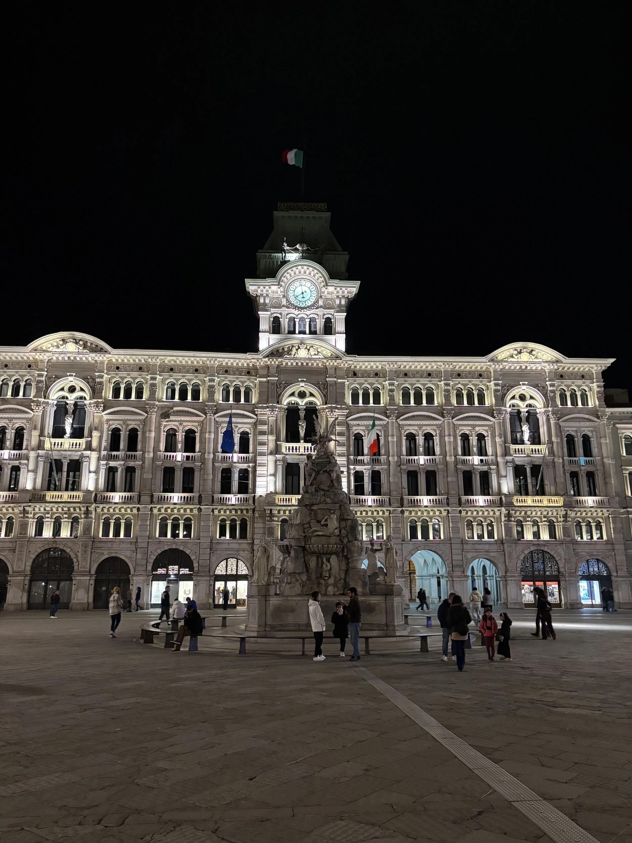 Night view of a historic illuminated building with a clock tower and Italian flag, with people gathered around a monument in the square in front.