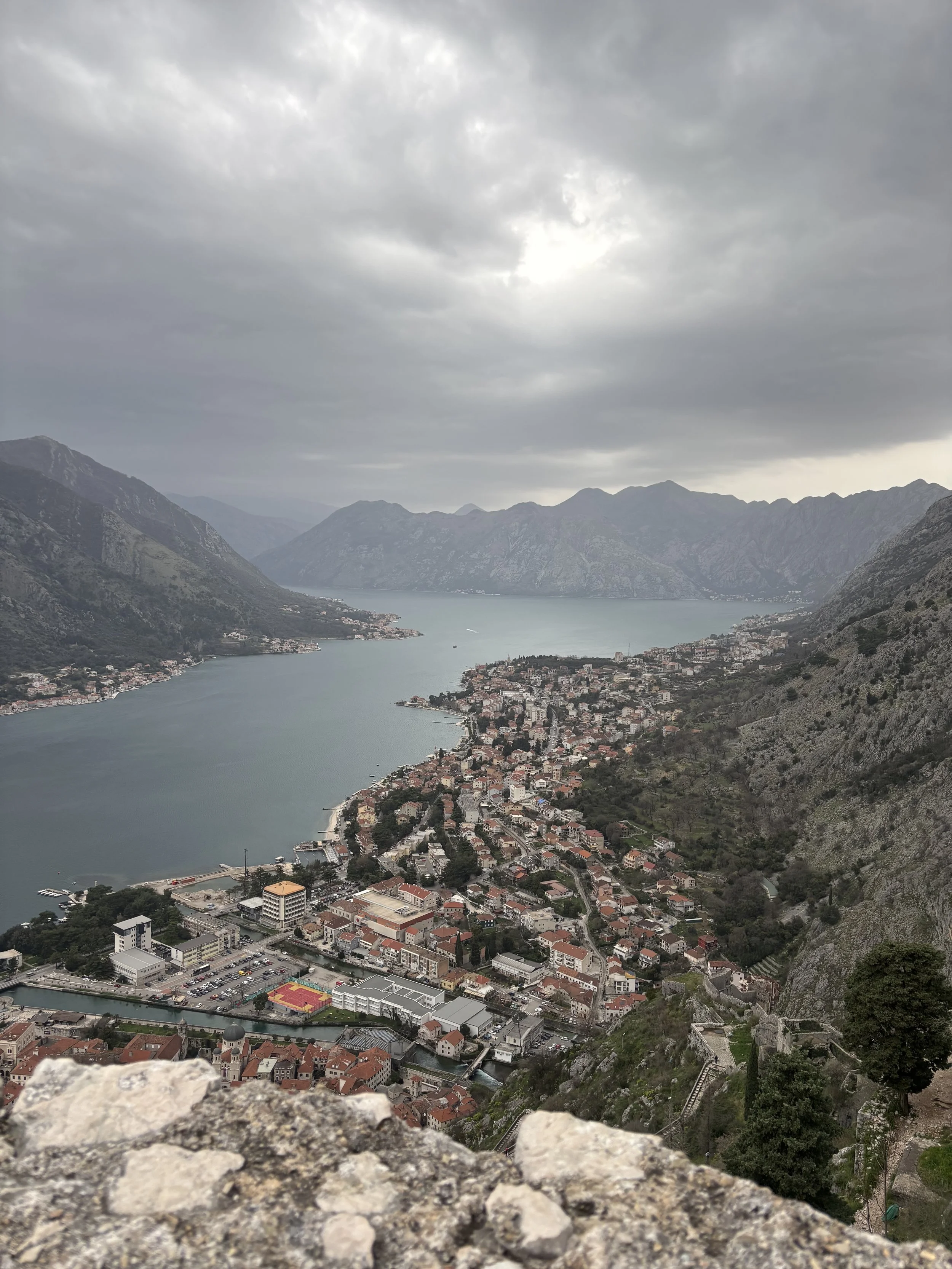 Aerial view of a coastal town nestled between mountains and a large body of water, under a cloudy sky.